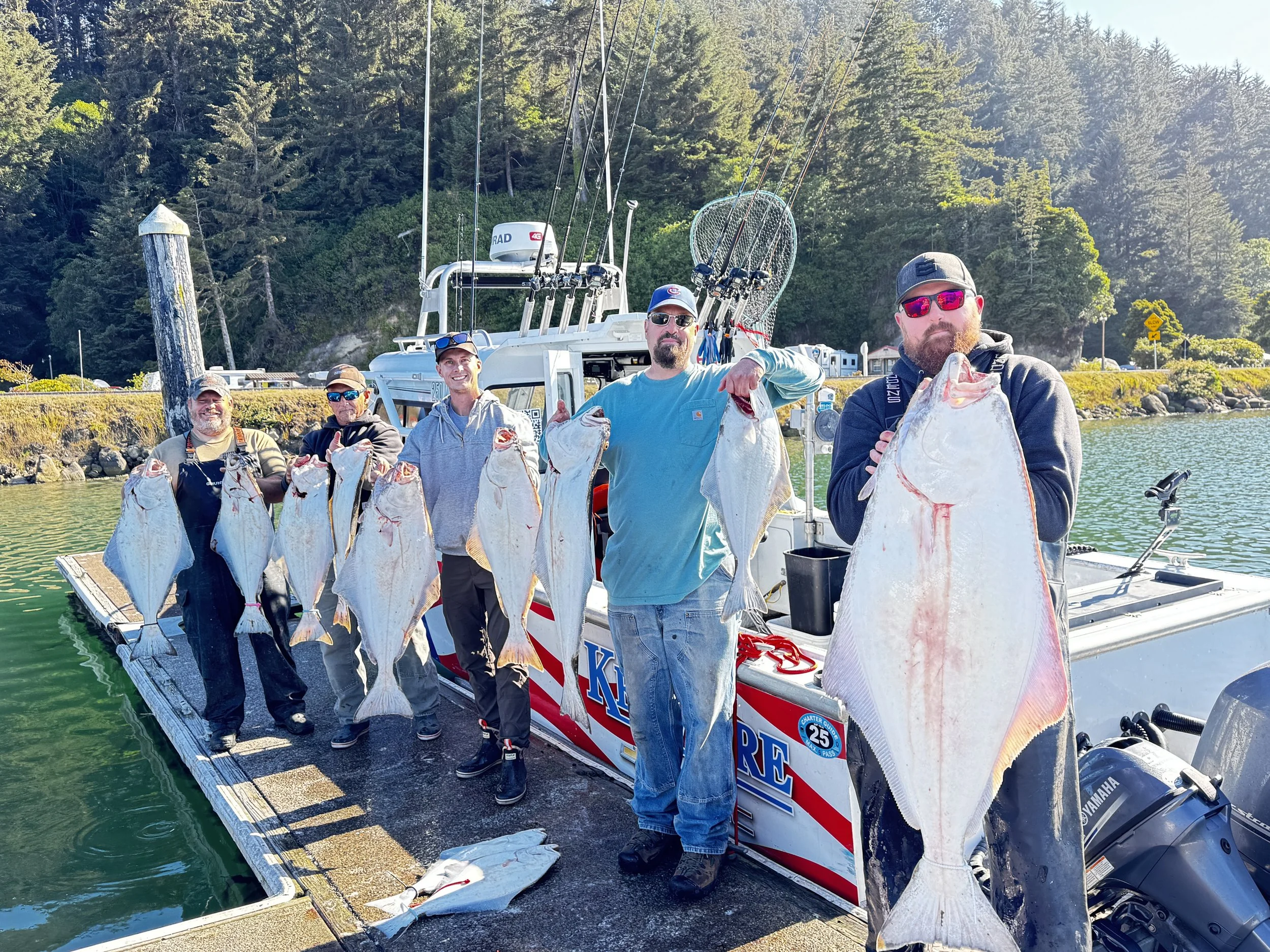 A group of five men holding fish on a dock at the water's edge with a boat, fishing gear, and a forested hillside in the background.