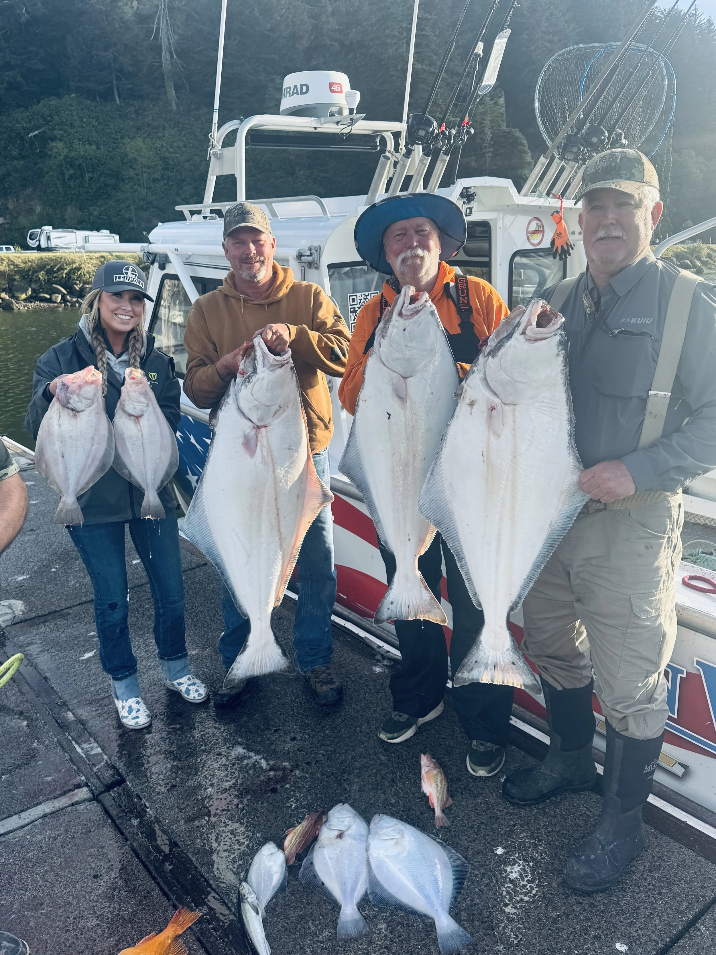 Four people standing on a dock in front of a boat, each holding a large fish they caught. The boat is equipped for fishing with fishing rods and a radar, and others are lying on the ground at their feet.