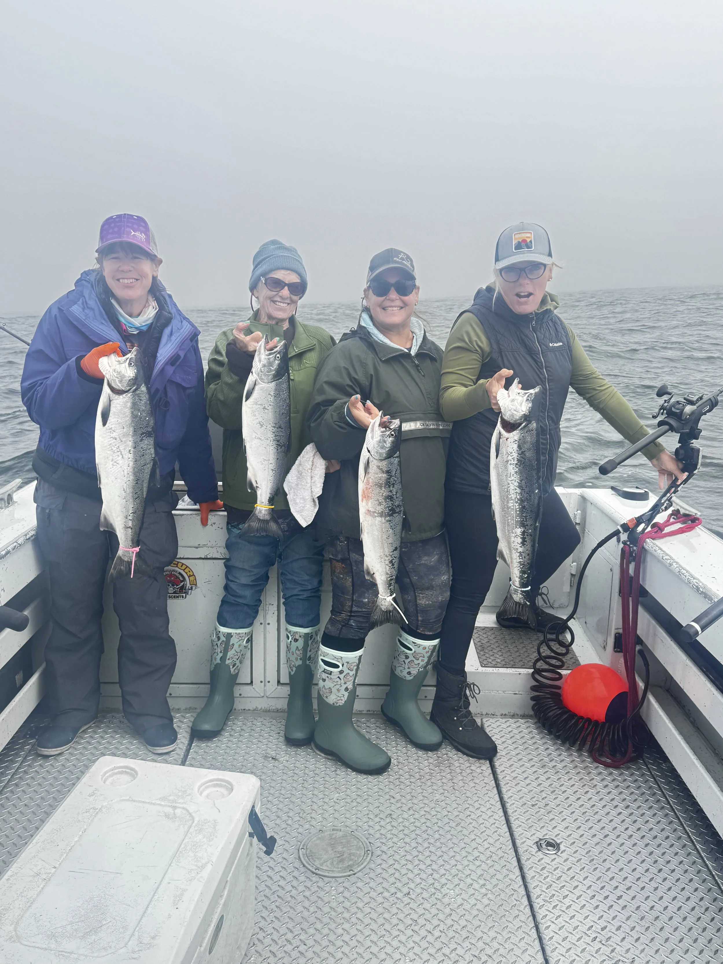 Four women are on a boat holding fish they caught, smiling, with a foggy ocean background.