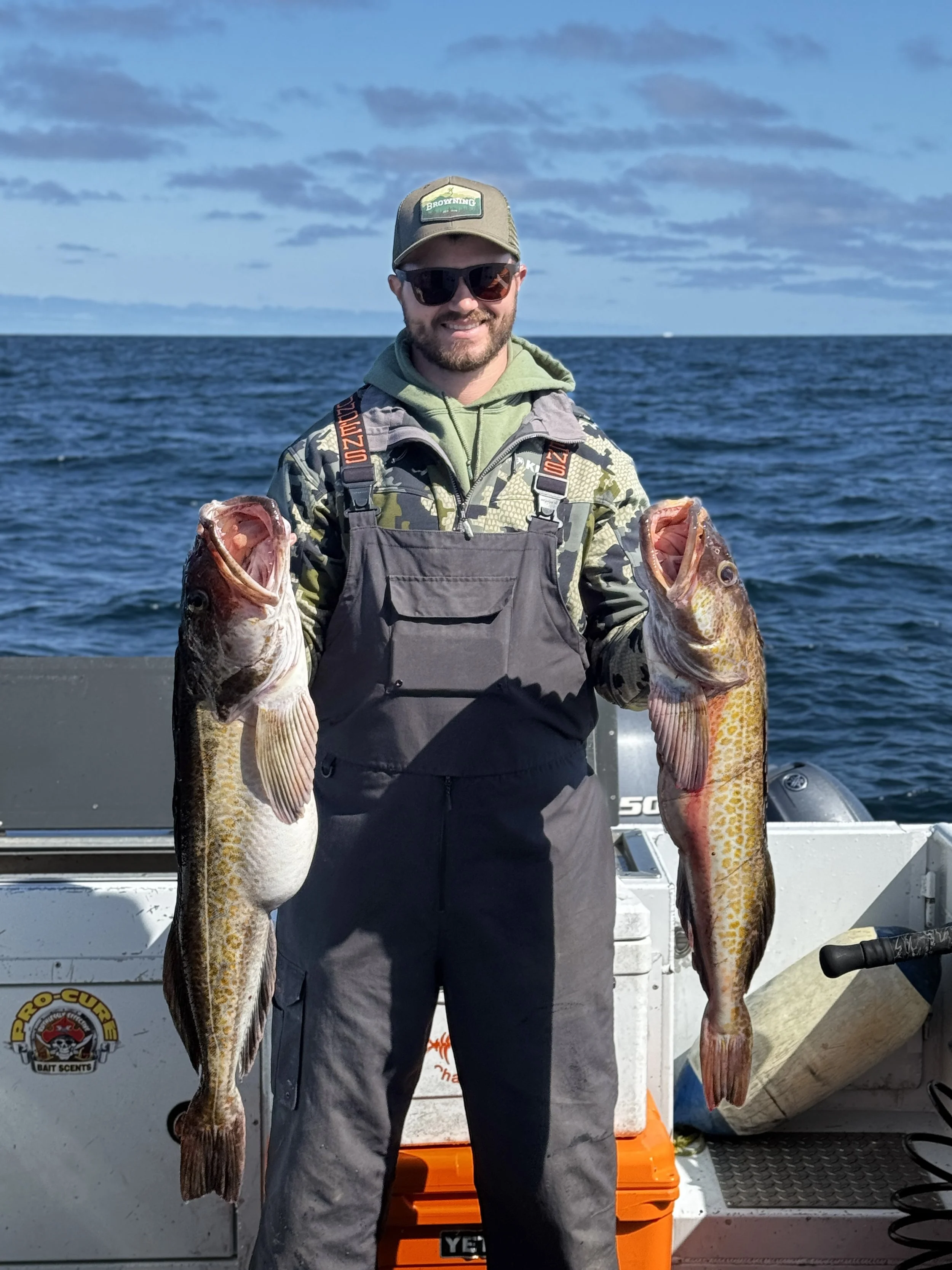 A man on a boat holding two large fish, smiling and wearing sunglasses, a hat, camouflage jacket, and overalls with a cloudy sky and ocean in the background.