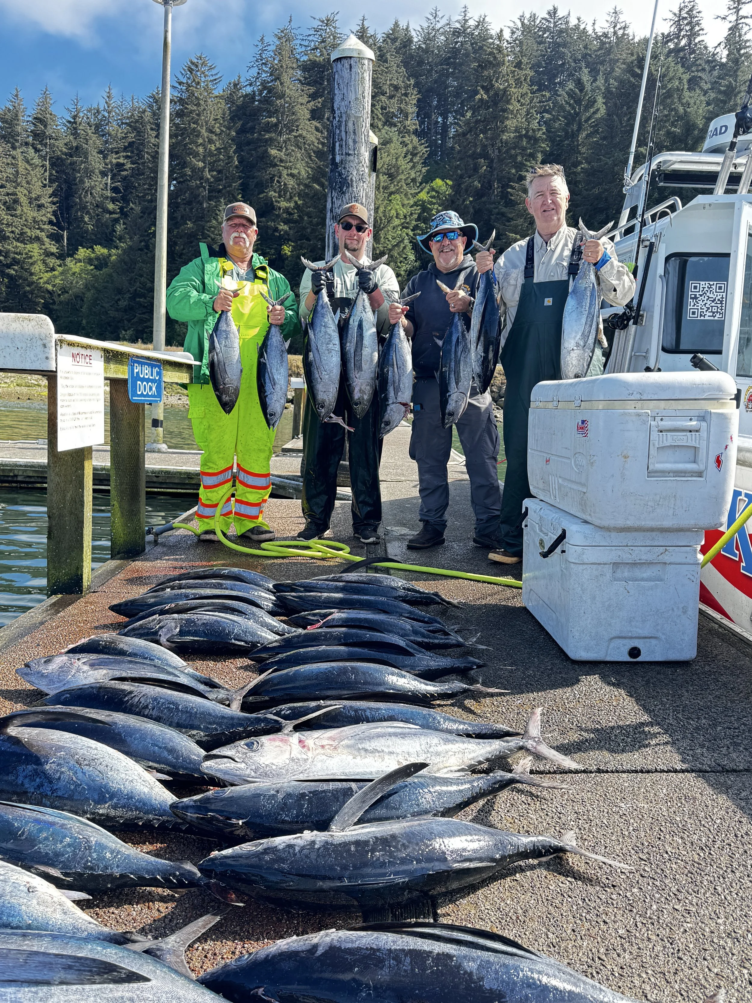 Four men standing on a dock holding fish with trees and a boat in the background, and fish laid out on the ground in front of them.