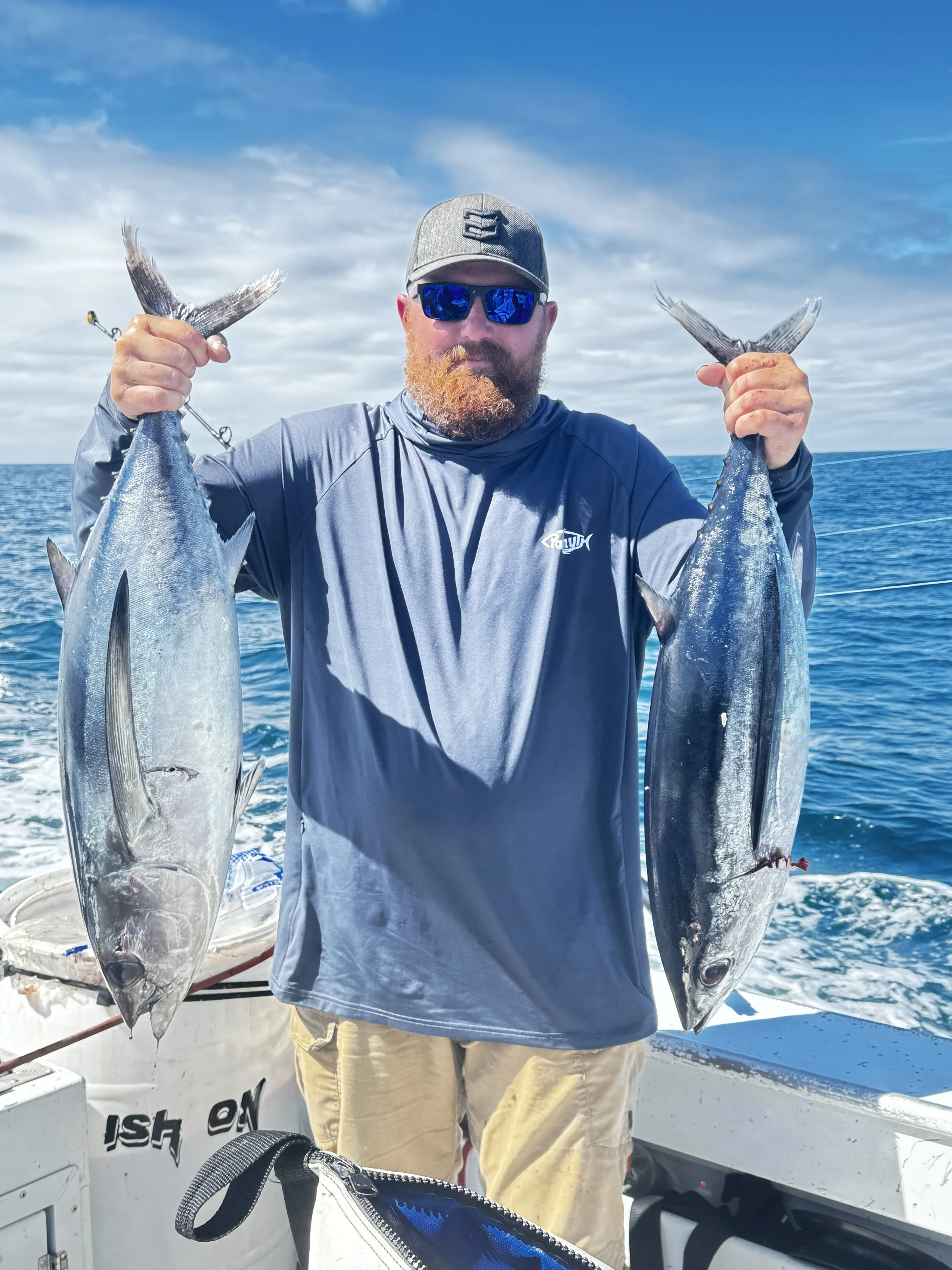 A man with a beard wearing sunglasses, a baseball cap, and a long-sleeve shirt is holding two large fish, one in each hand, on a boat with a view of the open ocean and a partly cloudy sky.