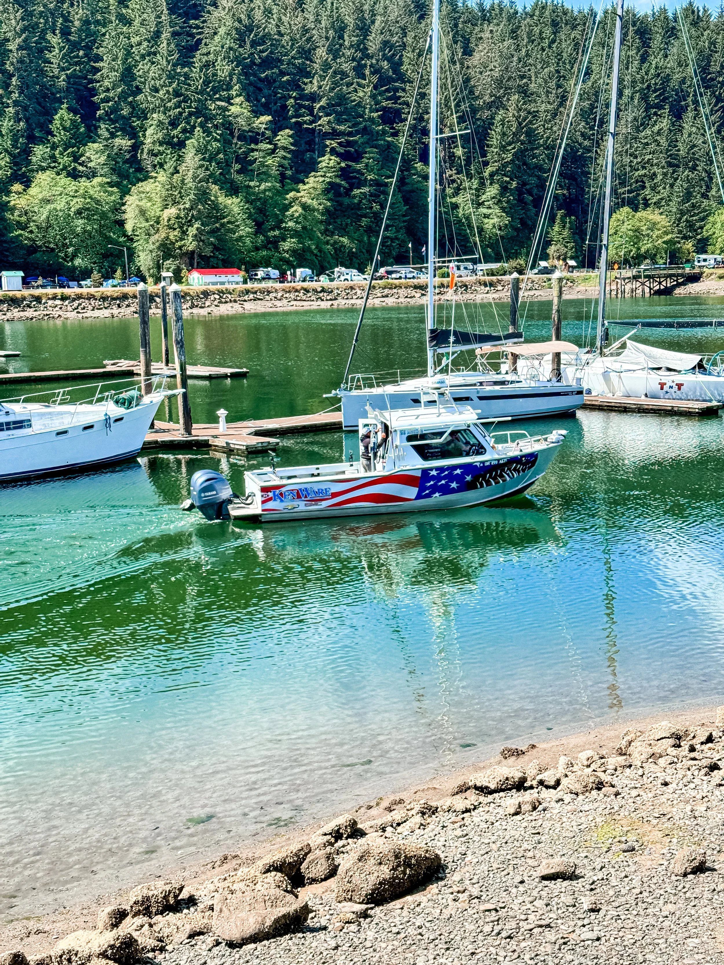 Boat with American flag design on a calm harbor, surrounded by sailboats and a forest background.