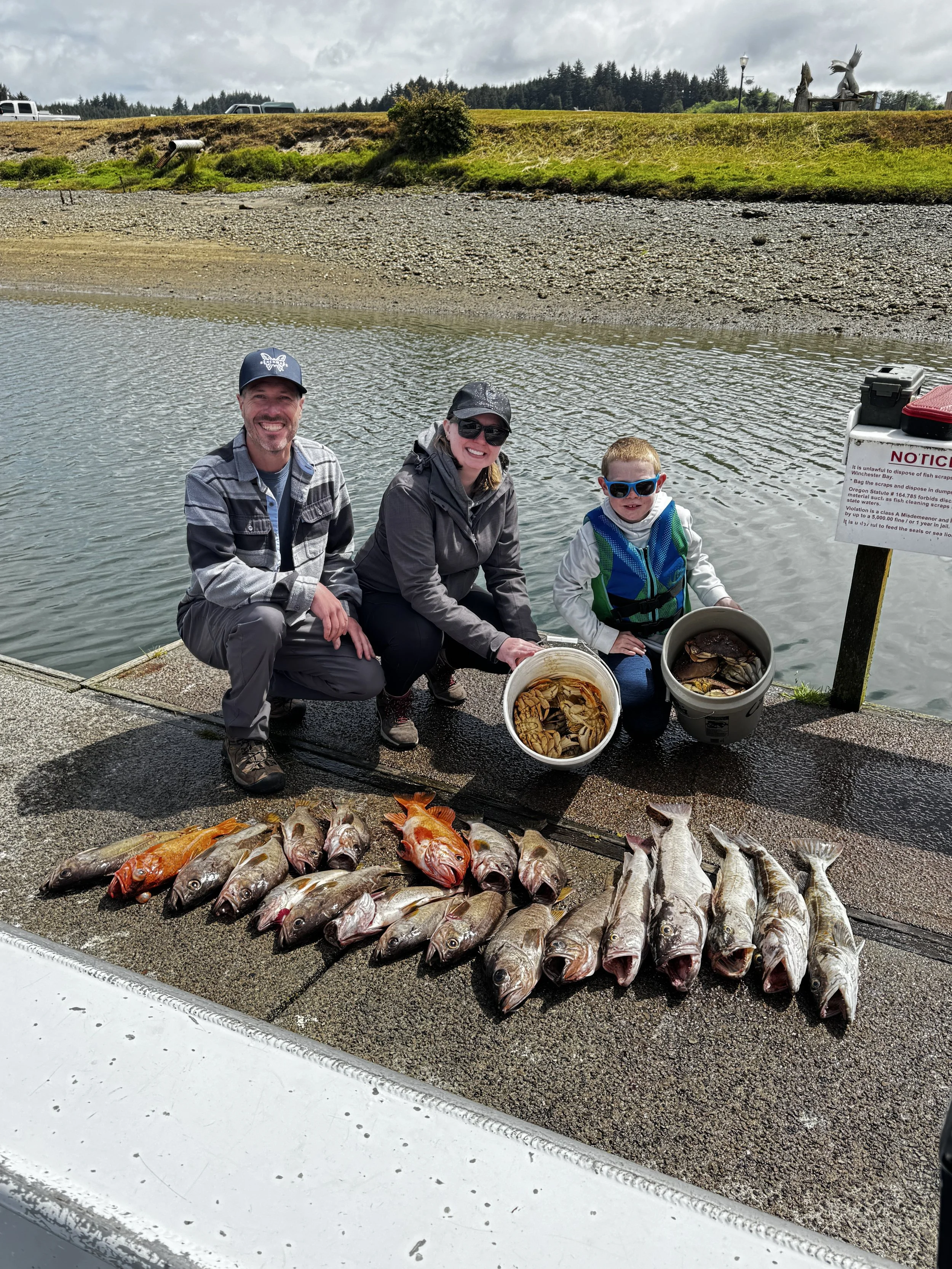Three people kneeling beside a catch of fish displayed on a dock, with buckets filled with fish, and a sign warning about fish disposal. The background features a grassy embankment and a cloudy sky.