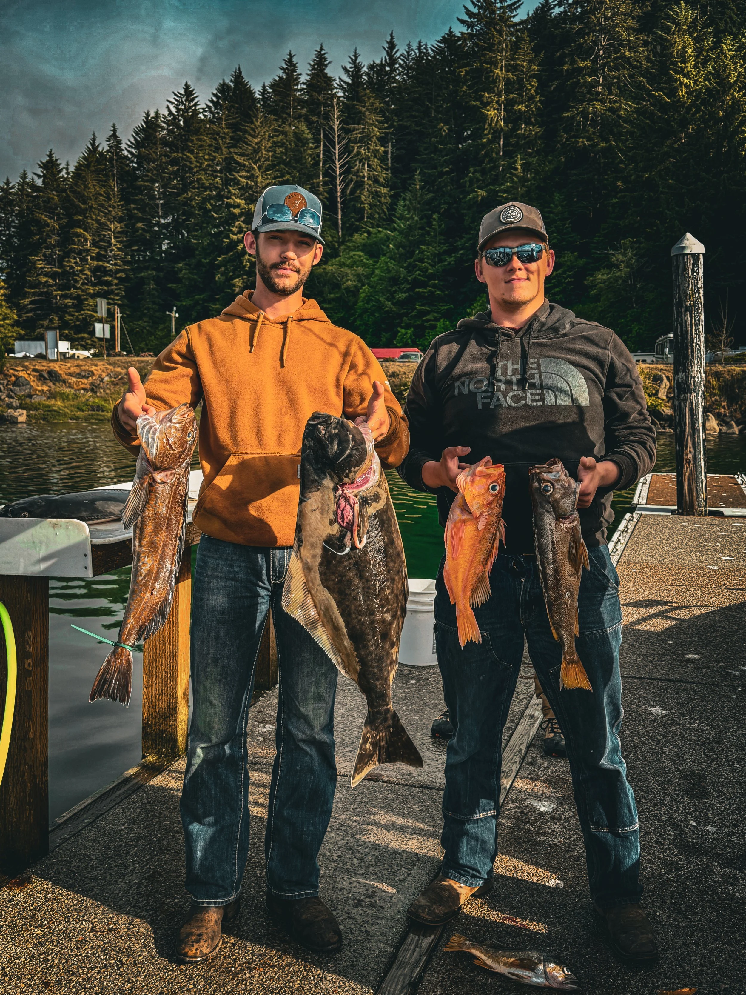 Two men standing on a dock holding various fish they caught, with a forested background.