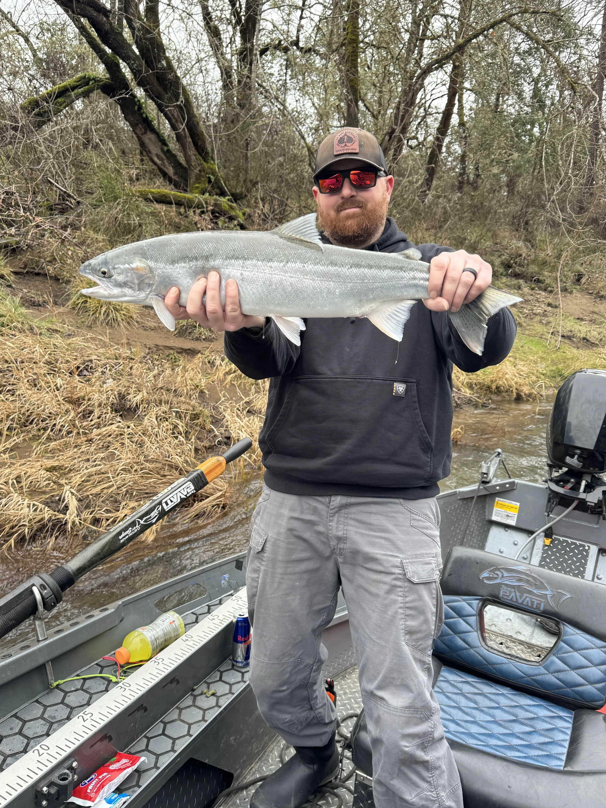 A man with a beard wearing a cap, sunglasses, black hoodie, and gray pants is standing in a boat on a river, holding a large fish with silver scales.