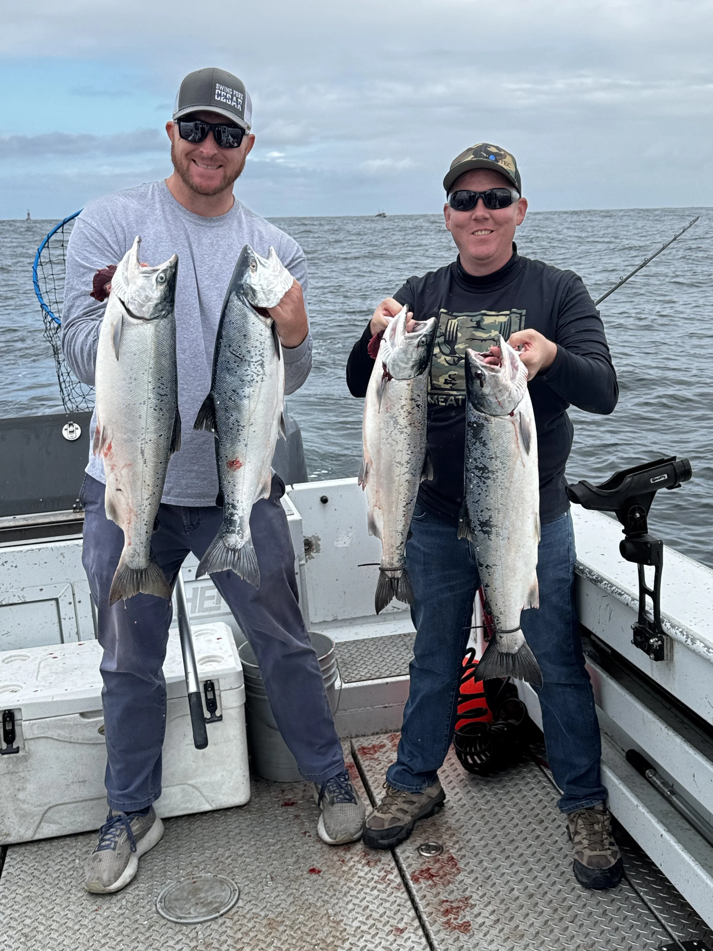Two men on a boat holding large fish, smiling, with an ocean background and cloudy sky.