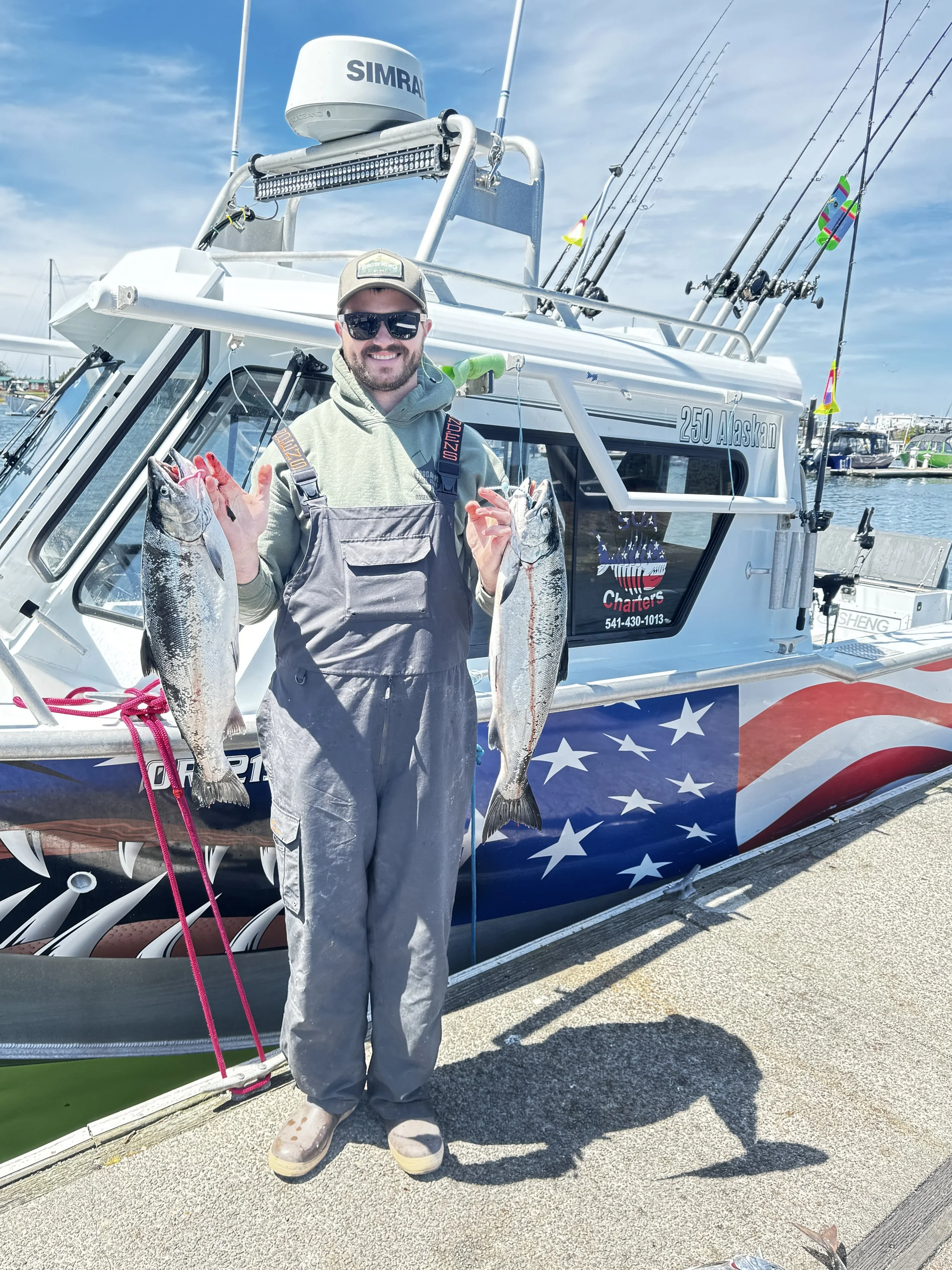 A man smiling and wearing sunglasses, a cap, and waterproof overalls, holding two fish in each hand, standing beside a boat decorated with an American flag design, at a marina with other boats docked in the background.