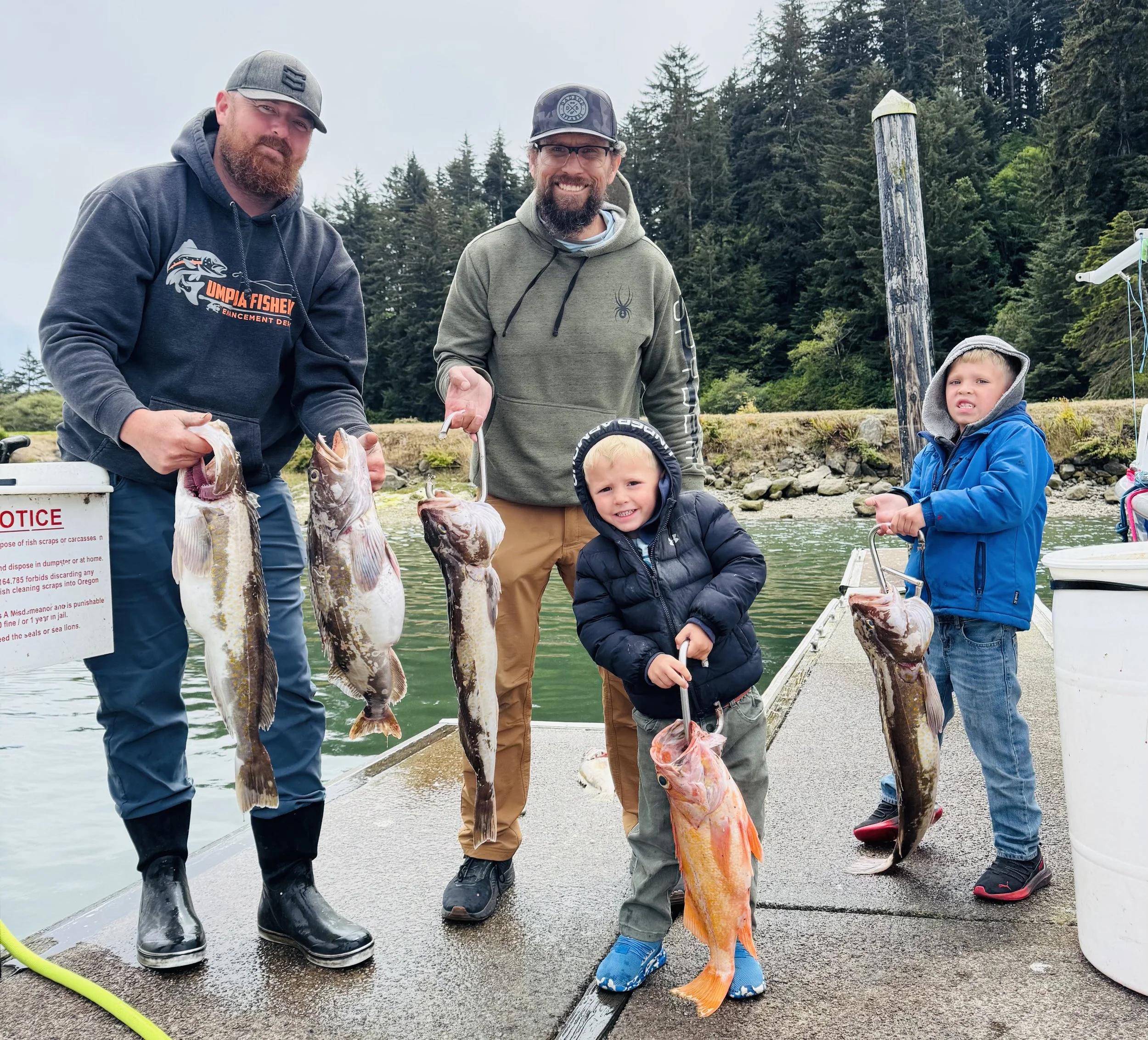 Two men and two boys holding large fish on a dock with a forest and water in the background.