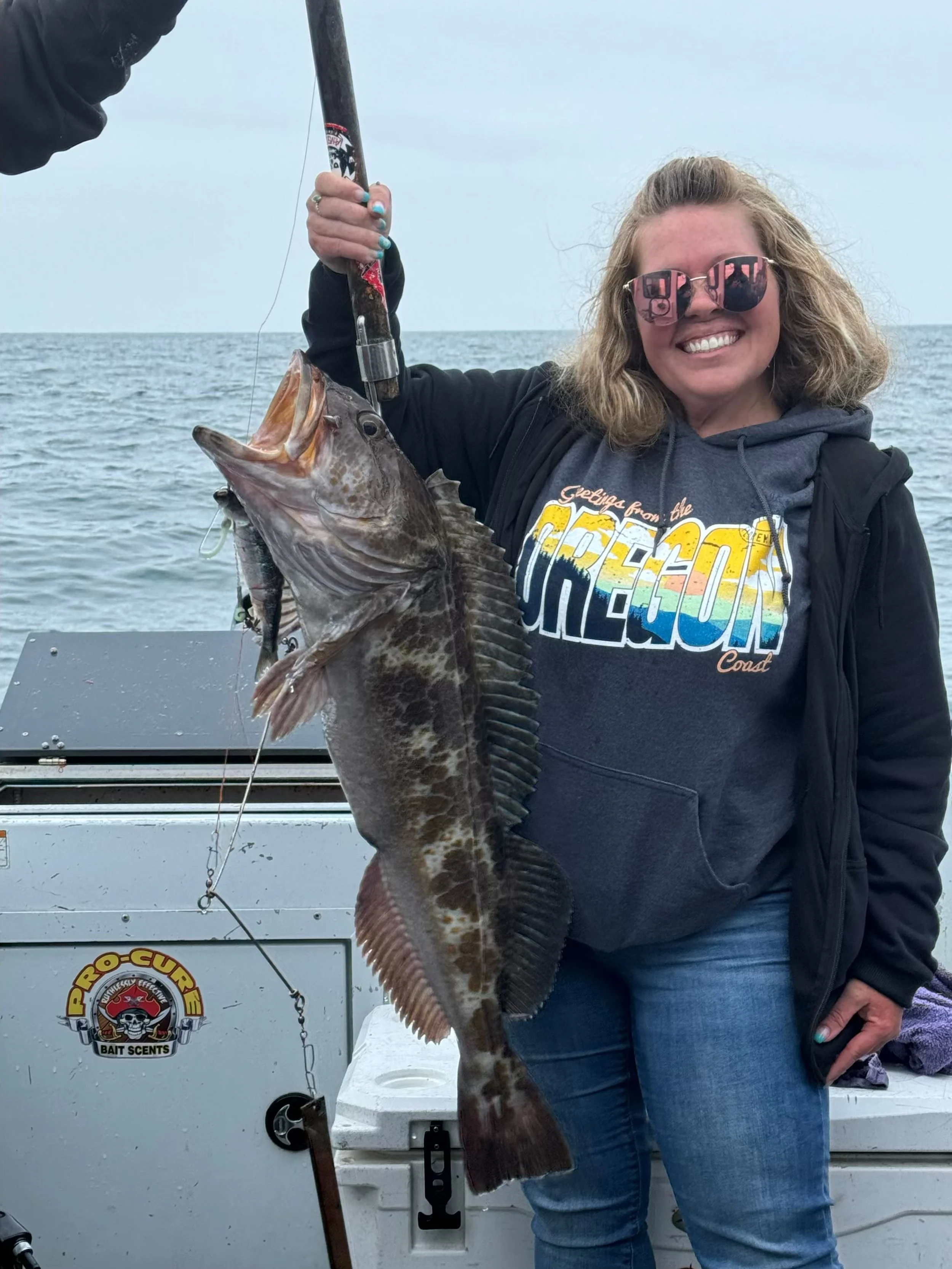 Woman holding a large fish on a boat, wearing sunglasses and an Oregon Coast sweatshirt, with ocean background.