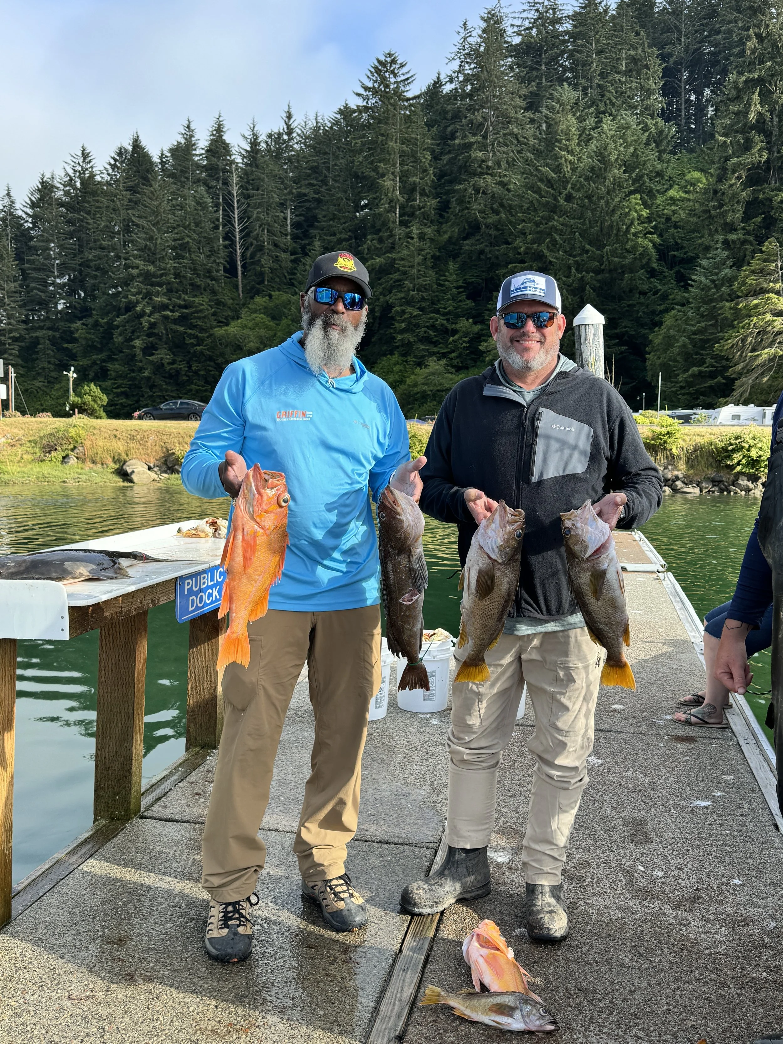Two men standing on a dock holding freshly caught fish, with a forested area in the background.