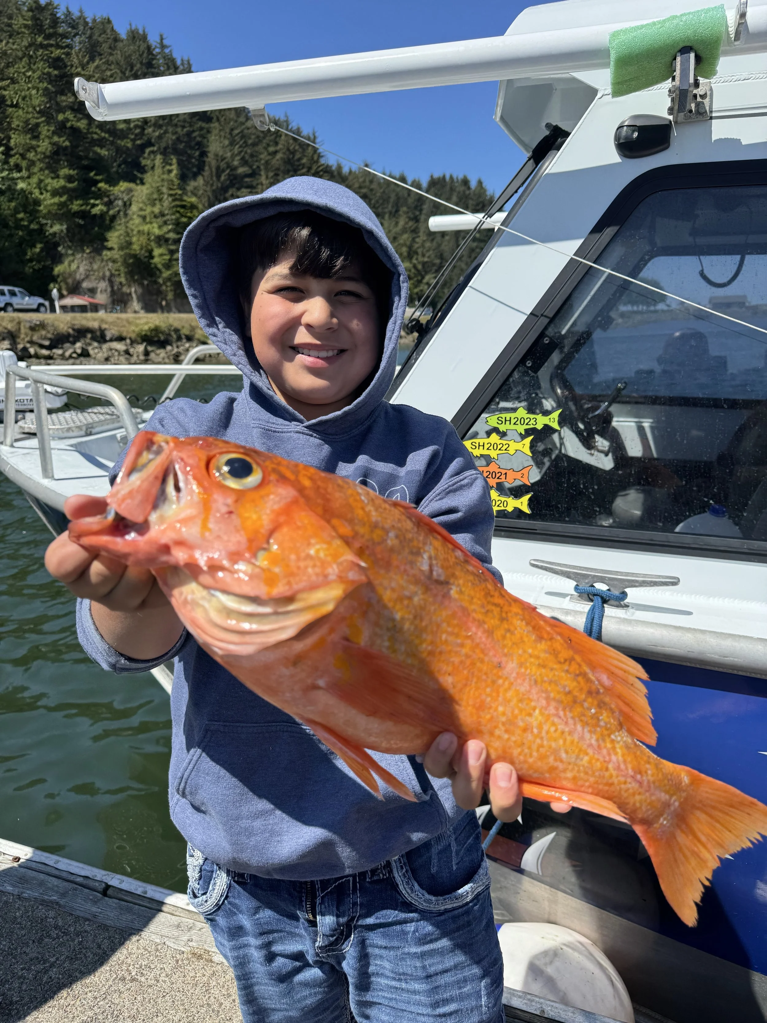 A young boy in a hoodie holding a large, colorful fish with orange and yellow scales on a boat dock.