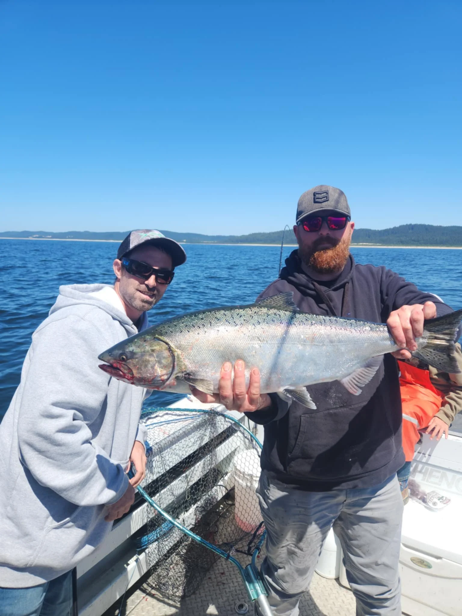 Two men are on a boat holding a large fish. They are wearing sunglasses and casual clothing, with a blue sky and water in the background.