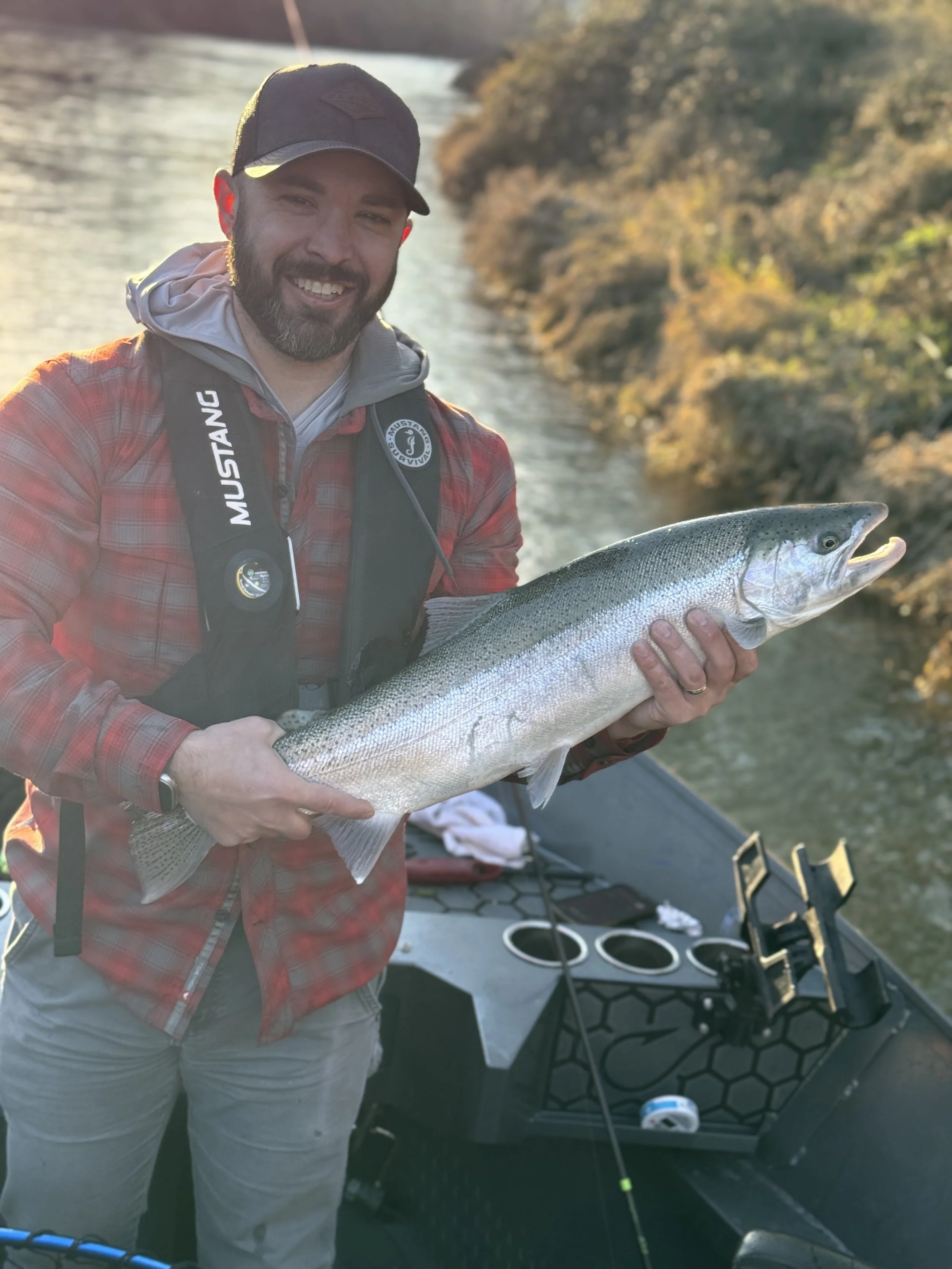A man with a beard smiling and holding a large fish, standing outdoors by a river at sunset.
