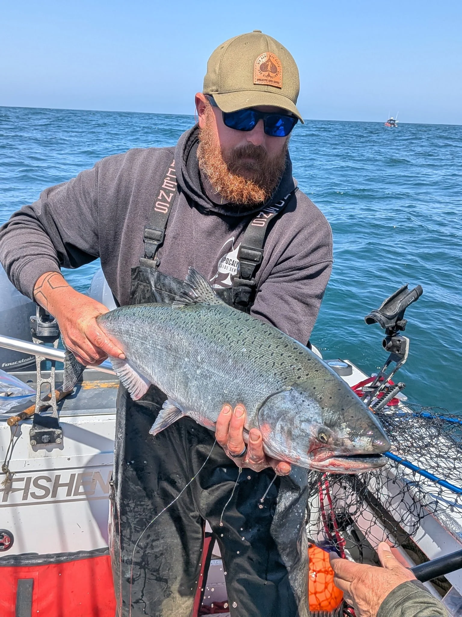 Man holding large fish on a boat at sea, wearing sunglasses, cap, and fishing gear, with fishing equipment and a boat visible in the background.