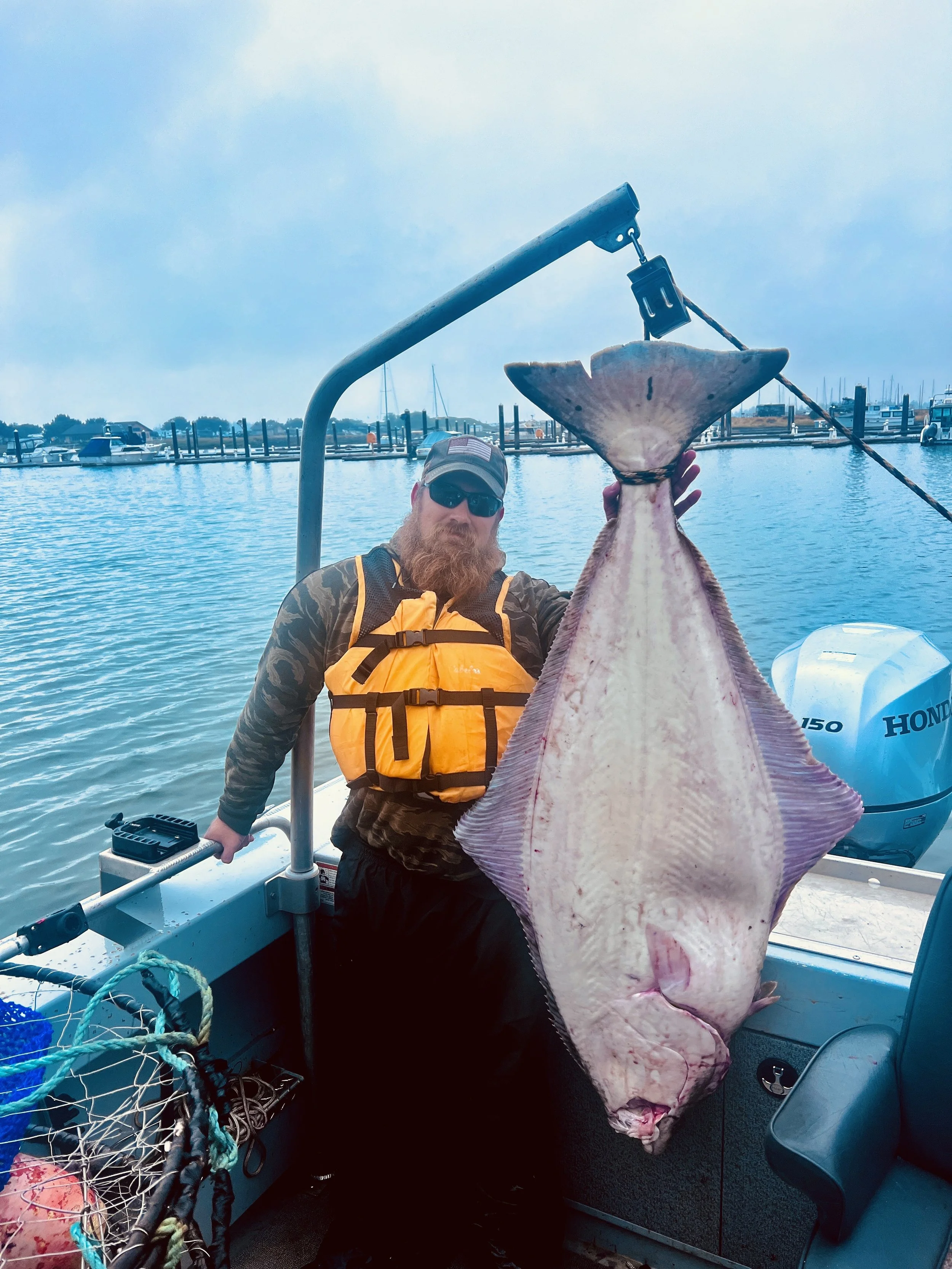 Fisherman holding large halibut on a boat with water and harbor in the background.