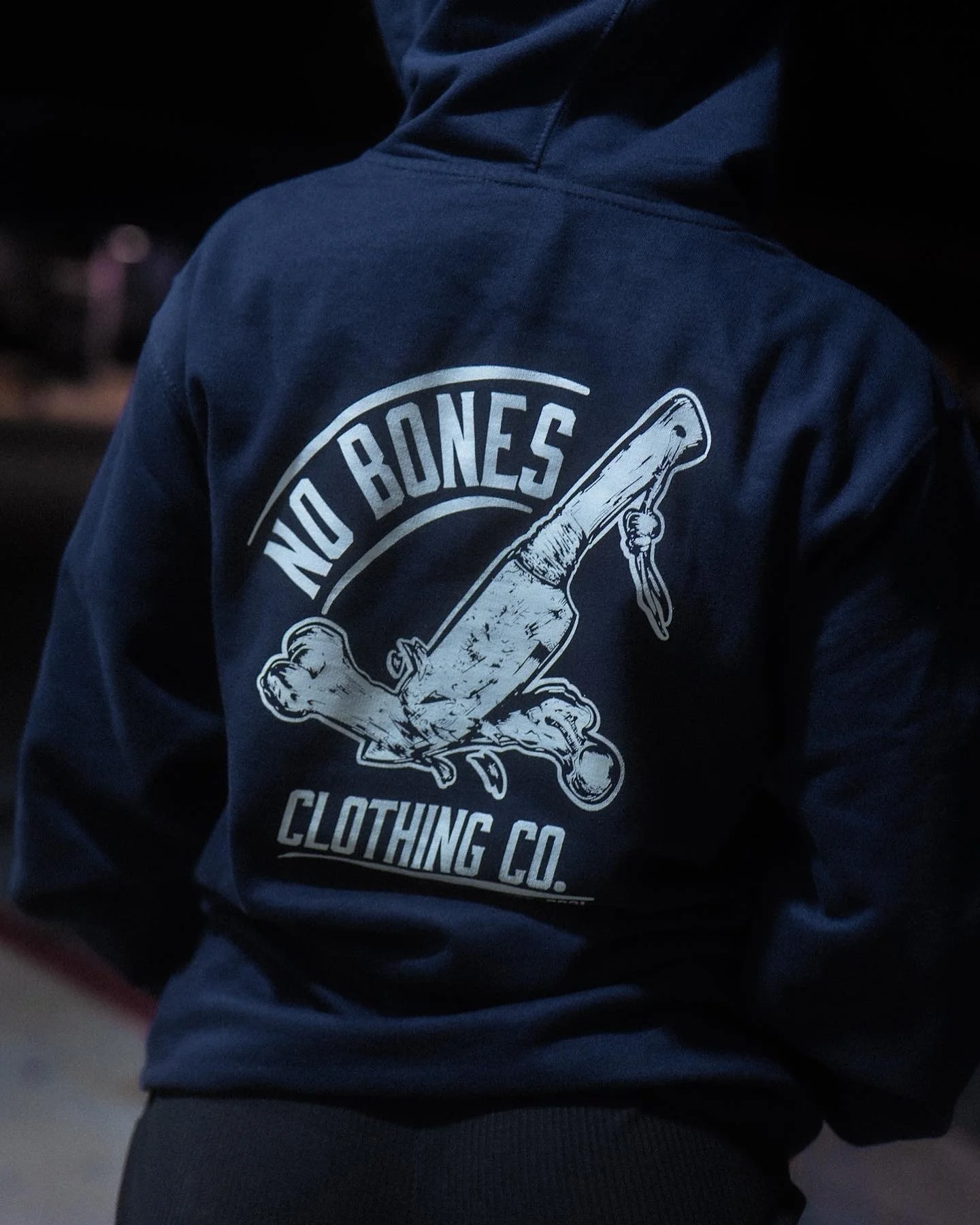 Person wearing a navy hoodie with a graphic and text that reads 'NO BONES CLOTHING CO', featuring a cartoon-style animated bone with a knife.