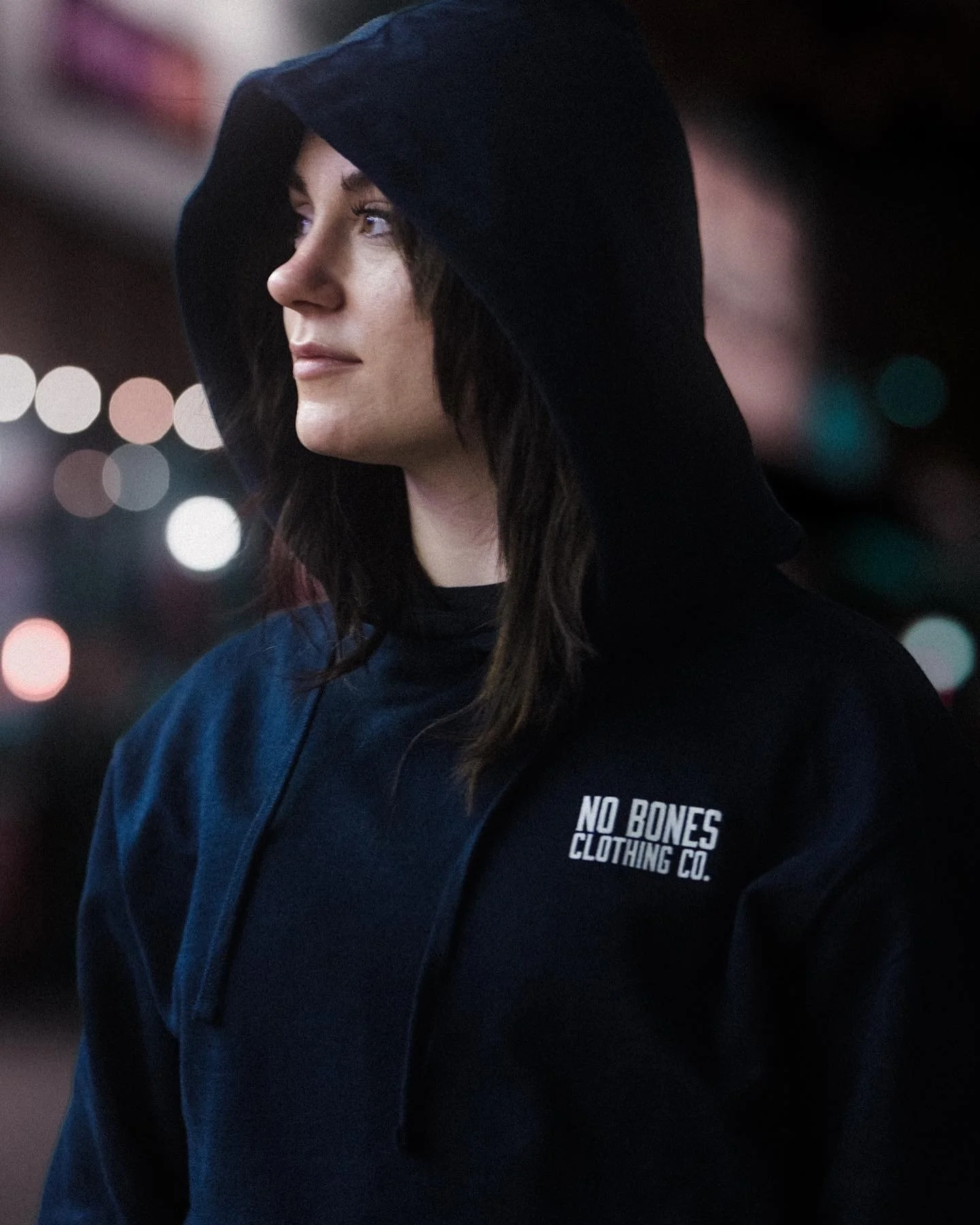 A woman with dark hair wearing a black hoodie with 'No Bones Clothing Co.' logo, standing outdoors at night with blurred city lights in the background.