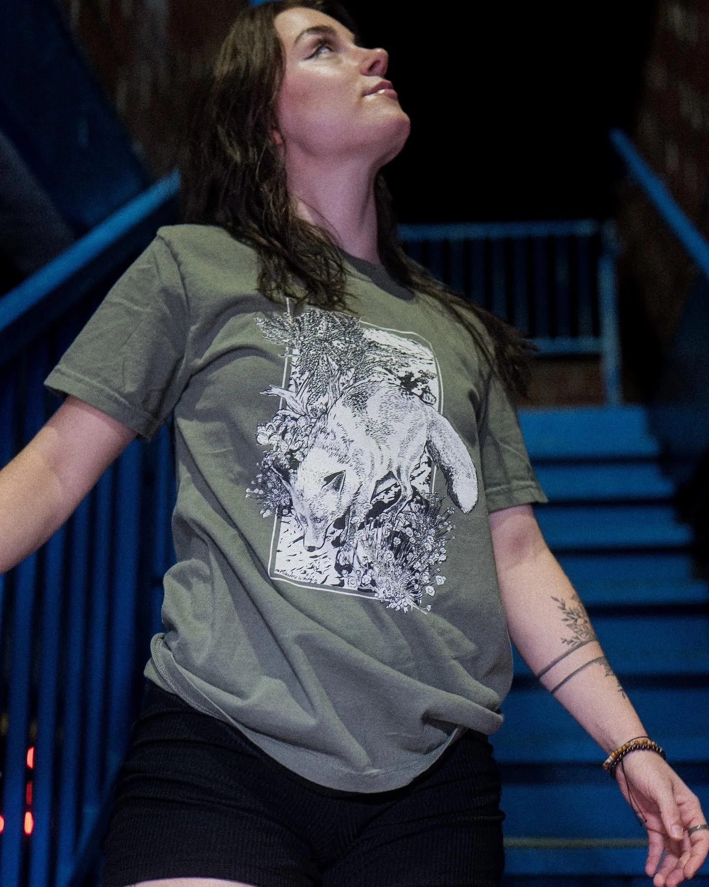 A woman with long brown hair, wearing a green T-shirt with an illustration of a fox and plants, stands on blue stairs looking upward.