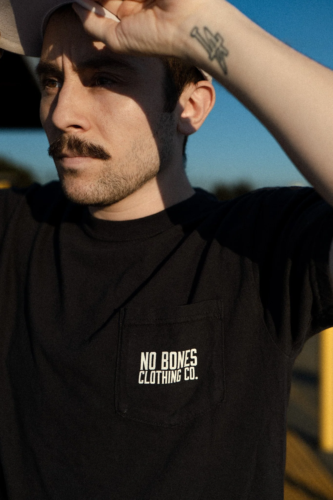 A man with a mustache and beard, wearing a black T-shirt with a pocket that reads 'NO BONES CLOTHING CO.' is outside during sunset, shielding his eyes with his hand.