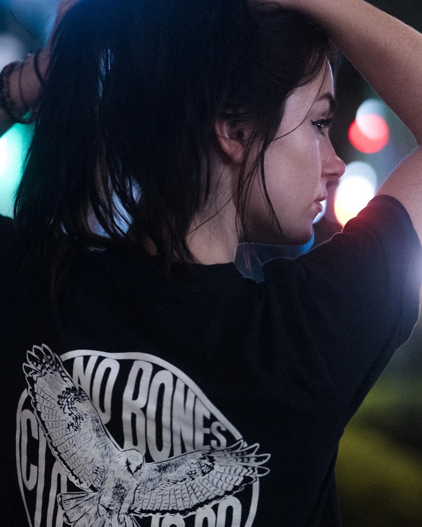 Side profile of a young woman with wet dark hair, wearing a black T-shirt with a white graphic of an owl and text, in a nighttime setting with blurred colorful lights in the background.