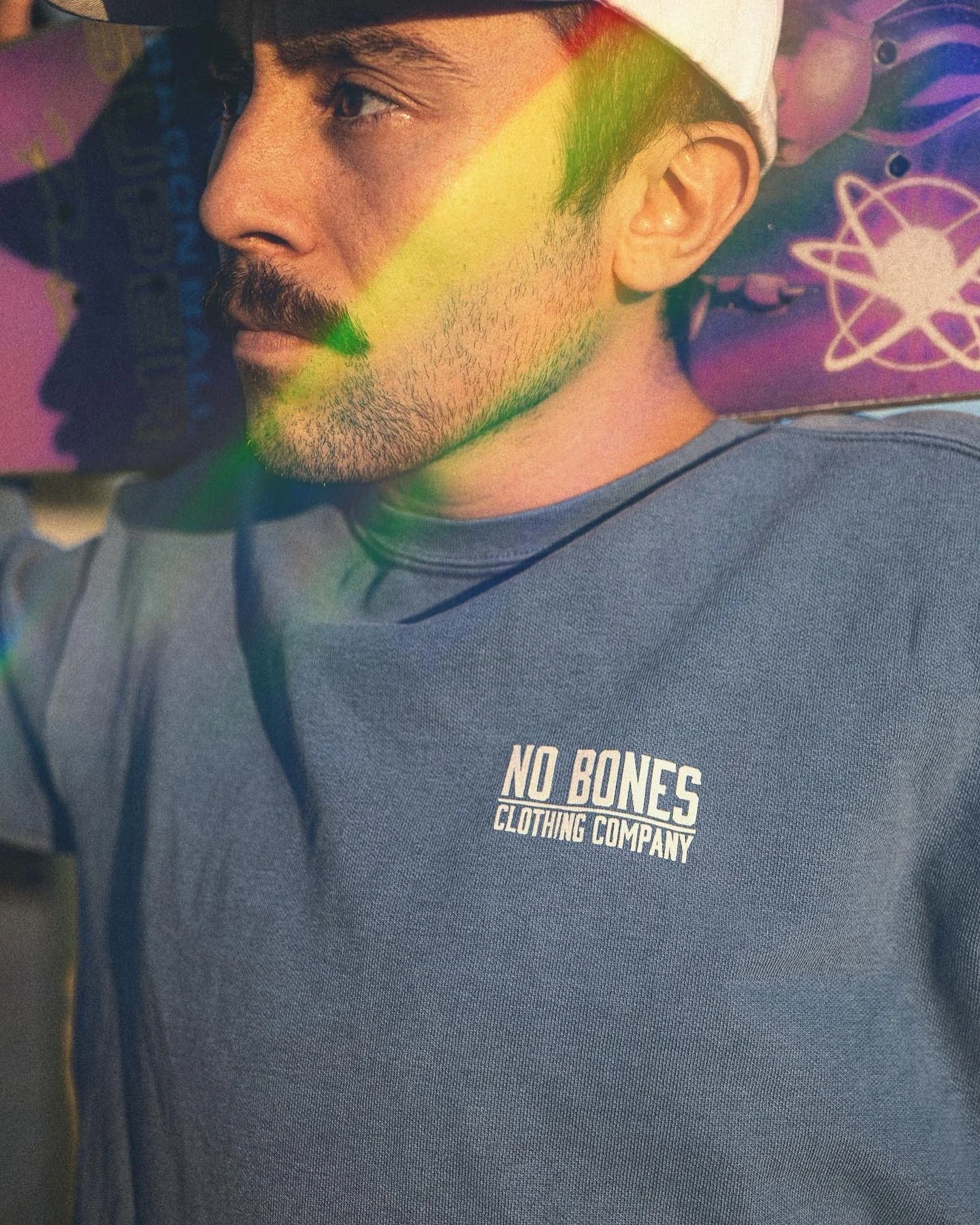 A man with a beard and mustache wearing a light-colored cap and a blue shirt with the text 'NO BONES CLOTHING COMPANY' on it, sitting against a colorful background with purple and pink hues and atom symbols.