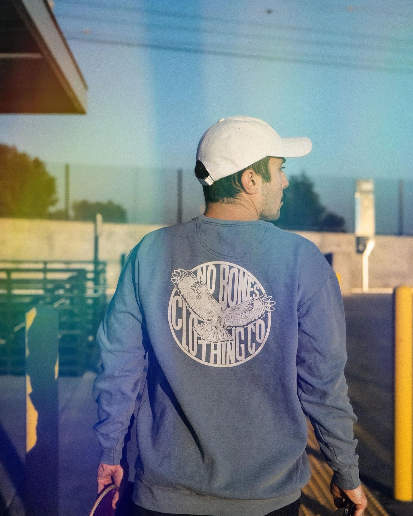 A man with dark hair wearing a white baseball cap and sweatshirt with a circular logo on the back showing an owl and text reading 'No Bones Gotta Thing Co.' walks down a sidewalk in an outdoor setting during dusk or dawn, holding a skateboard in his 