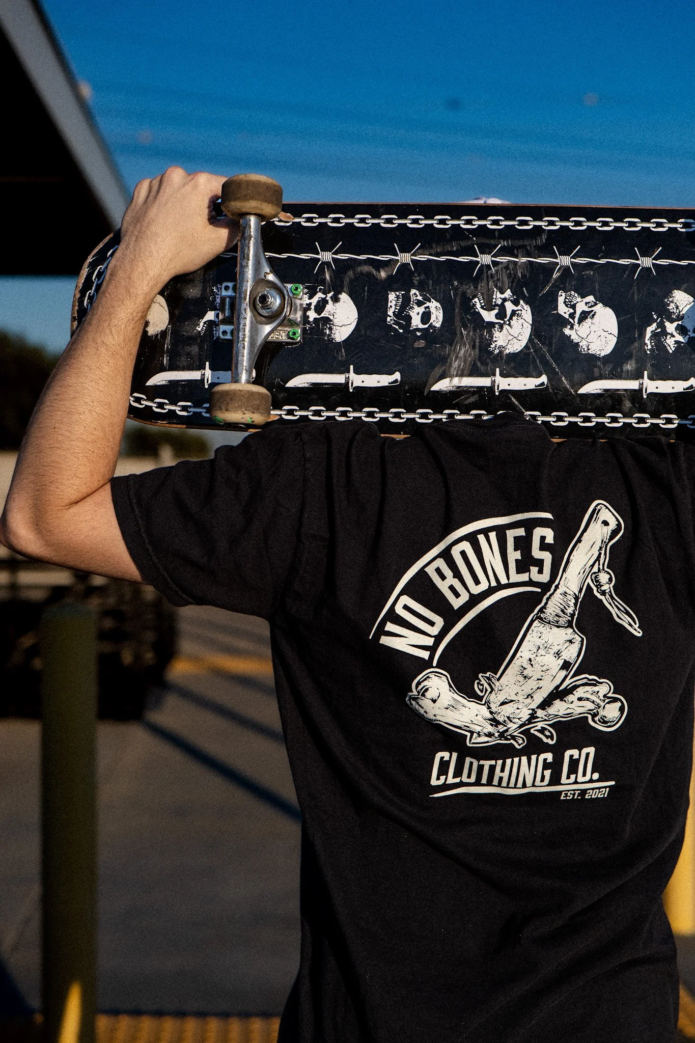 Person holding skateboard over shoulder, wearing a black T-shirt with a graphic of a bone and the words 'NO BONES CLOTHING CO.'