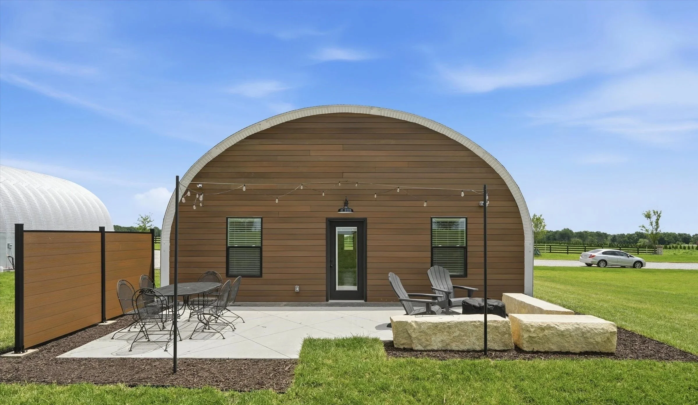 A small wooden Quonset hut-style building with a black door, two windows, and string lights hanging overhead. In front, there are patio chairs and a table, with a large stone and chairs nearby. A fenced grassy field, a silver car, and another curved building are visible in the background under a blue sky.
