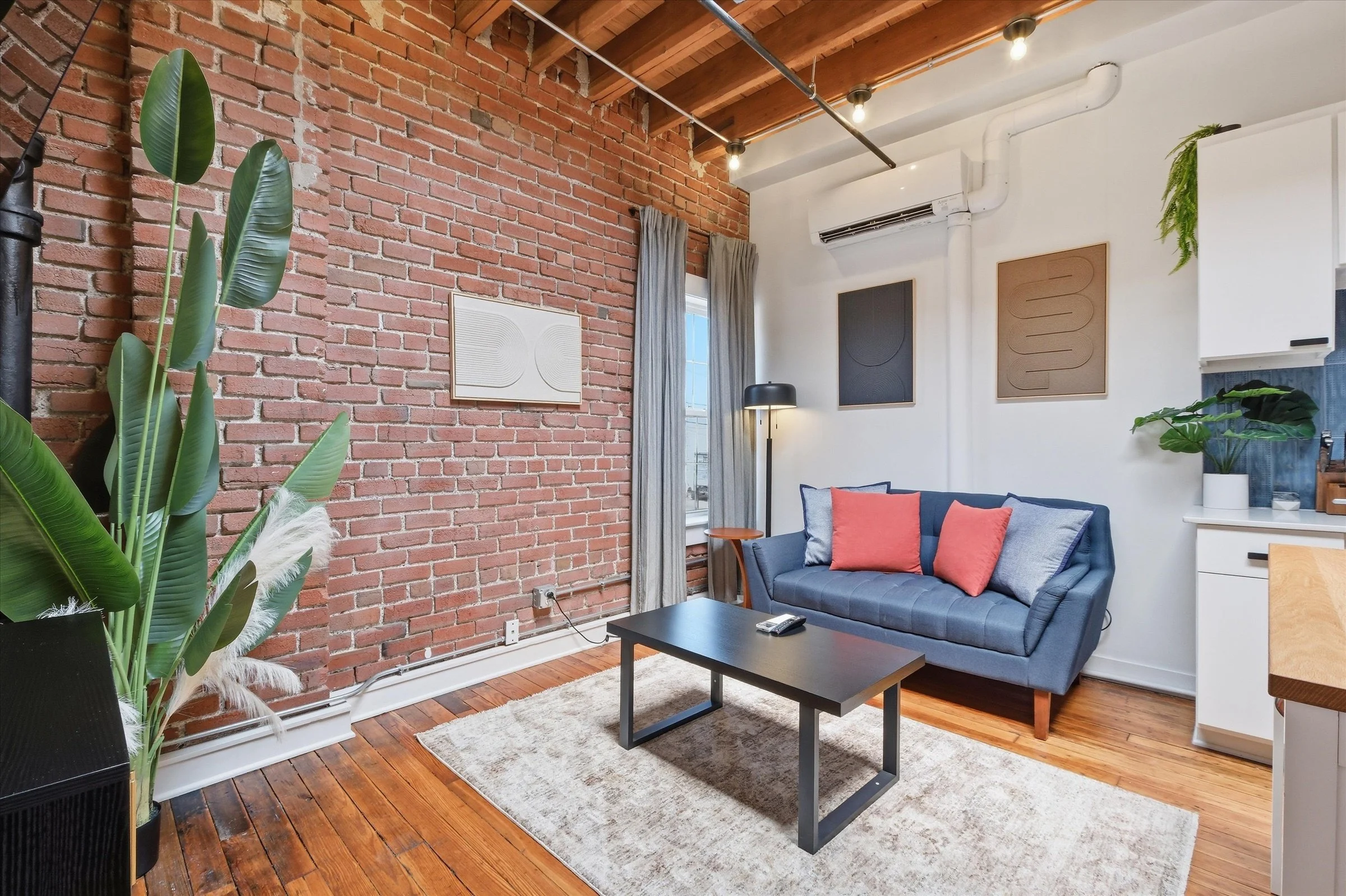 A cozy living room with an exposed red brick wall, wooden ceiling beams, and a white wall with black and brown abstract art. There is a blue sofa with orange and gray cushions, a black coffee table, a side table with a floor lamp, and large green indoor plants. A window with curtains allows natural light in.