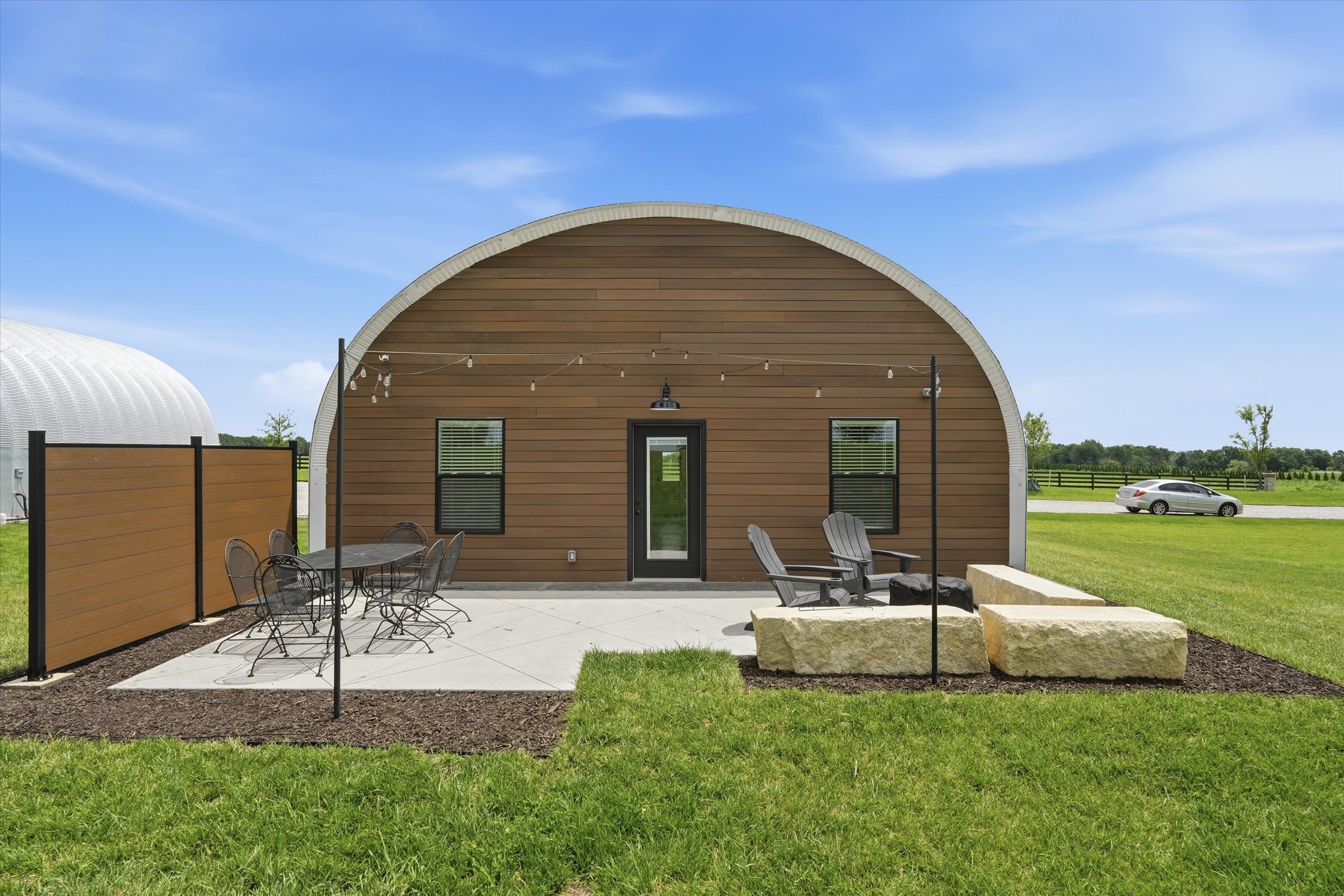 A modern arched wood-paneled building with outdoor seating and string lights, set in a grassy area under a blue sky.