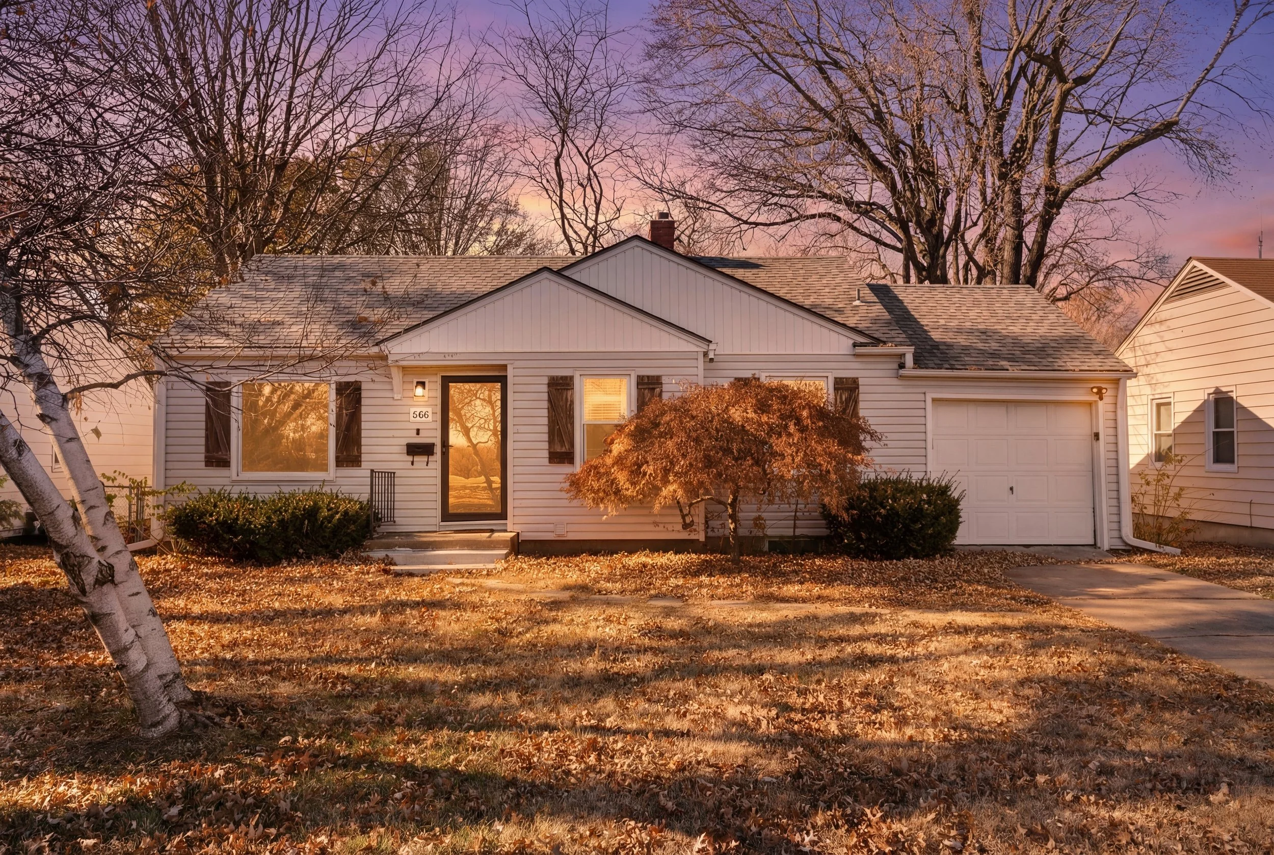A white suburban house with a garage, front door, and large windows, surrounded by autumn trees with fallen leaves on the lawn and sidewalk, under a colorful pink and purple sunset sky.