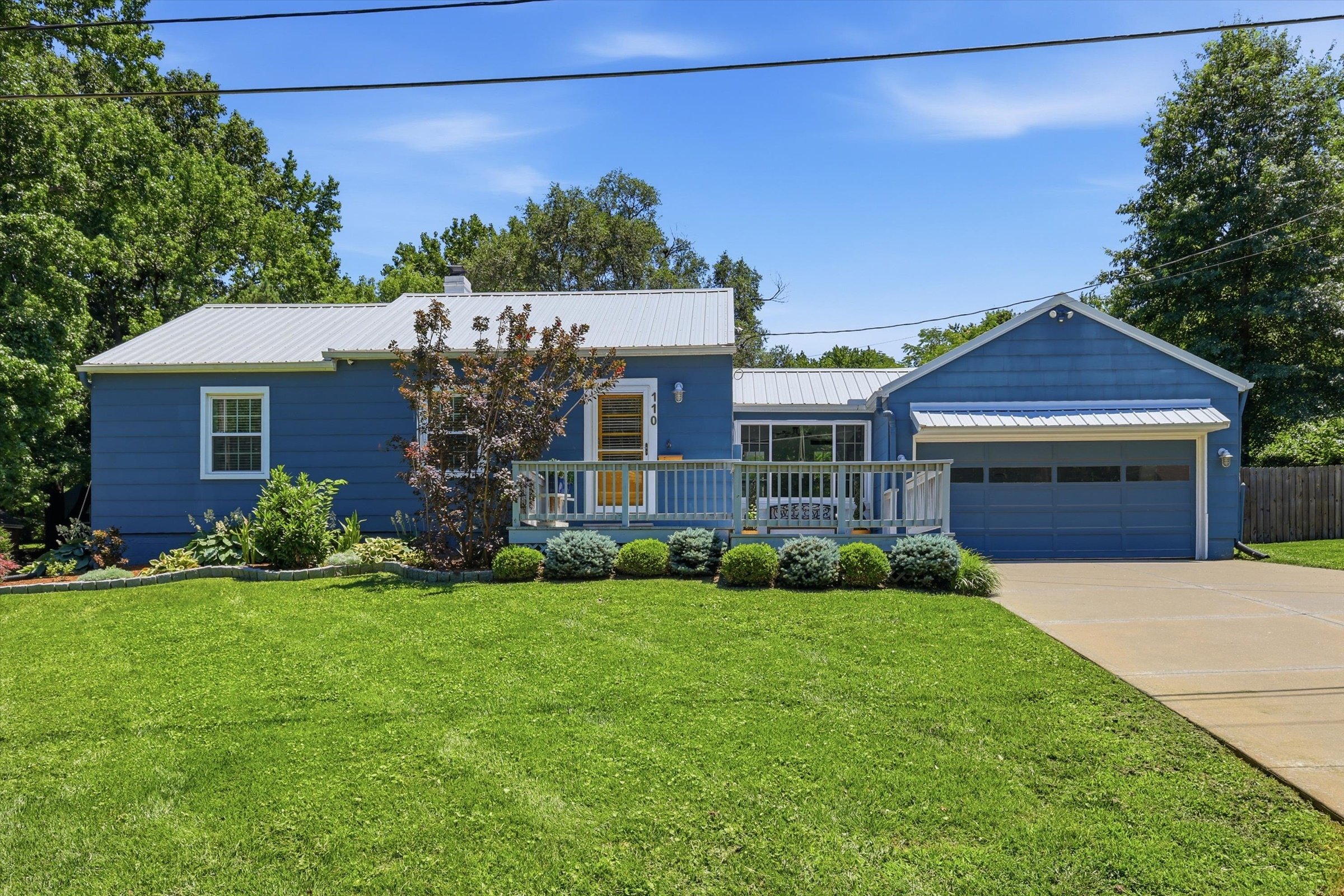 A blue house with a metal roof, front porch, and attached garage. Well-maintained green lawn with flower bed and small tree in front. Clear blue sky with some clouds, large trees behind the house, and power line overhead.