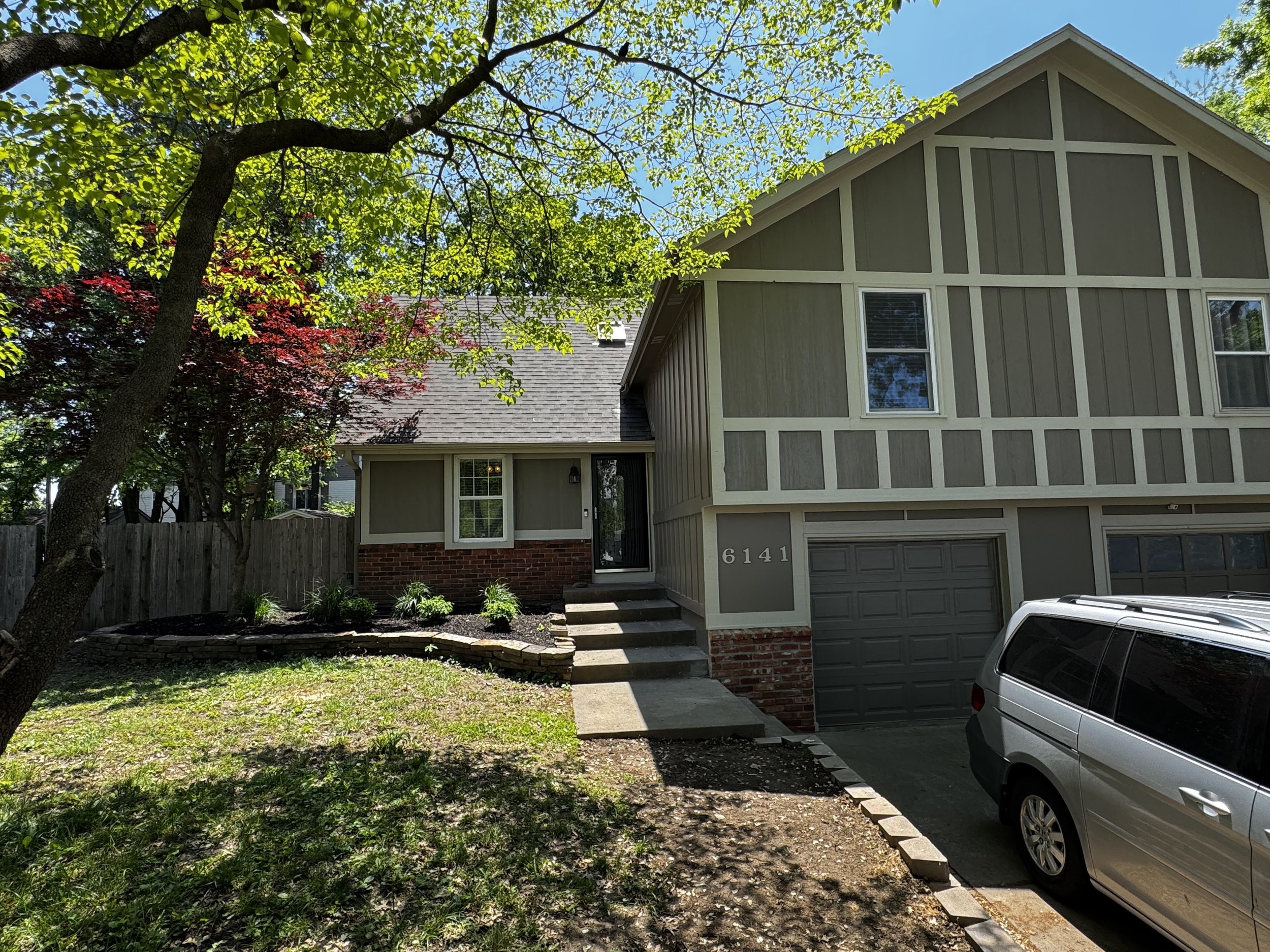 Front view of a two-story house with a gray exterior and white trim, surrounded by green trees and a small front yard with grass and plants, parked car in driveway.