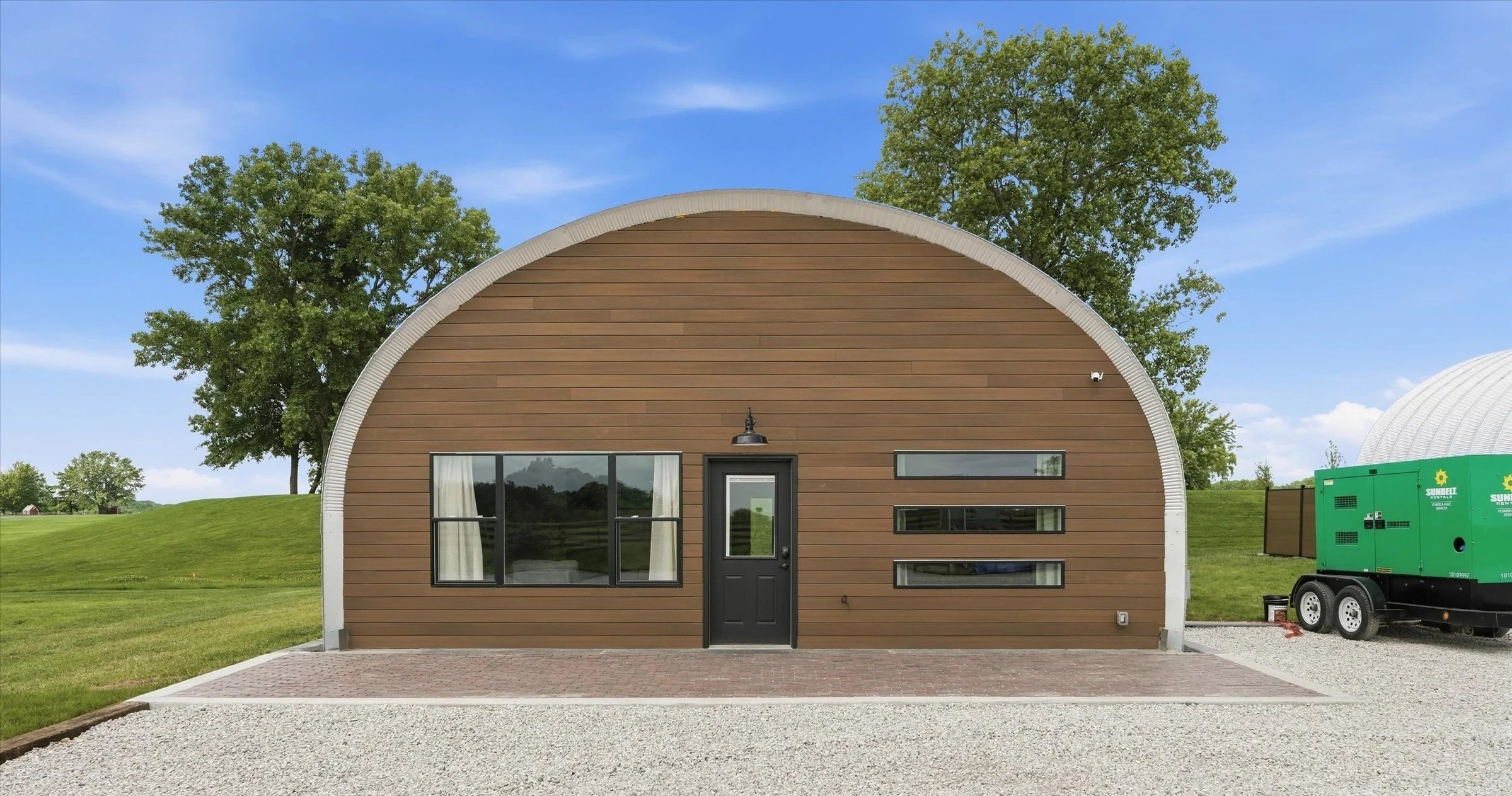 A modern gable-roof tiny house with brown horizontal siding, black door, and horizontal windows, set on a grassy landscape with trees and a clear blue sky.