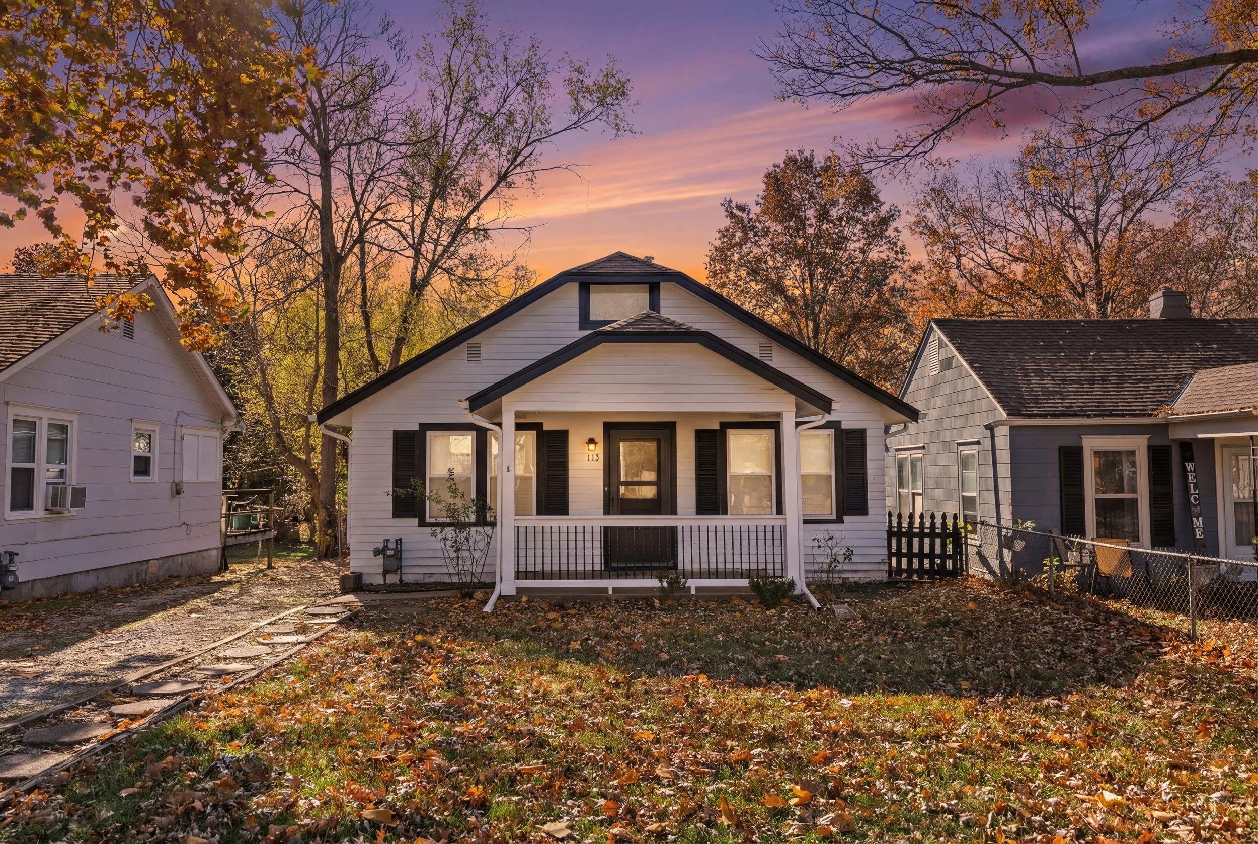 A small white house with black trim at sunset, surrounded by trees with autumn leaves and empty yard.
