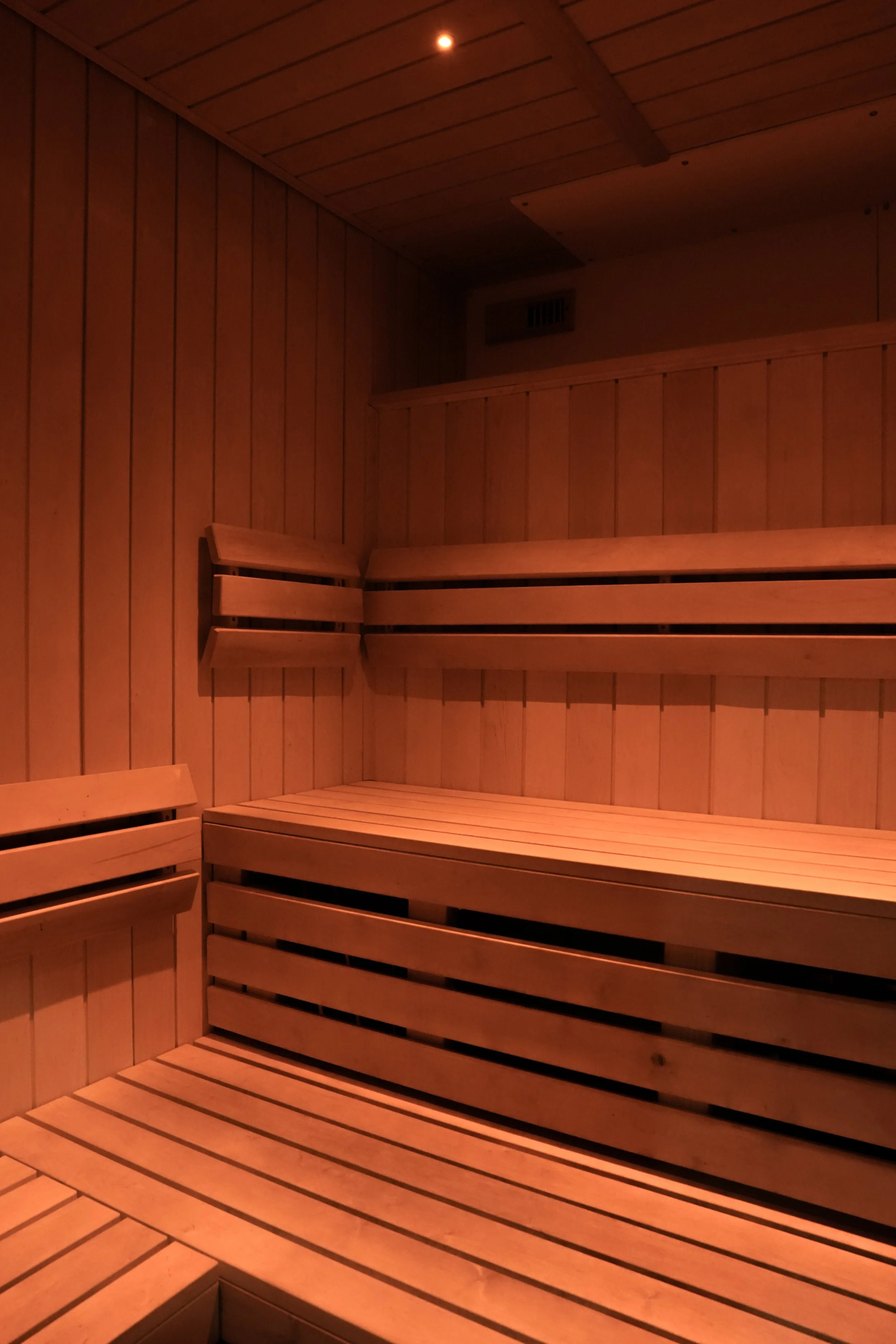 Interior of a modern wooden sauna featuring multi-level bench seating, warm ambient lighting, and smooth vertical wood paneling on the walls and ceiling.