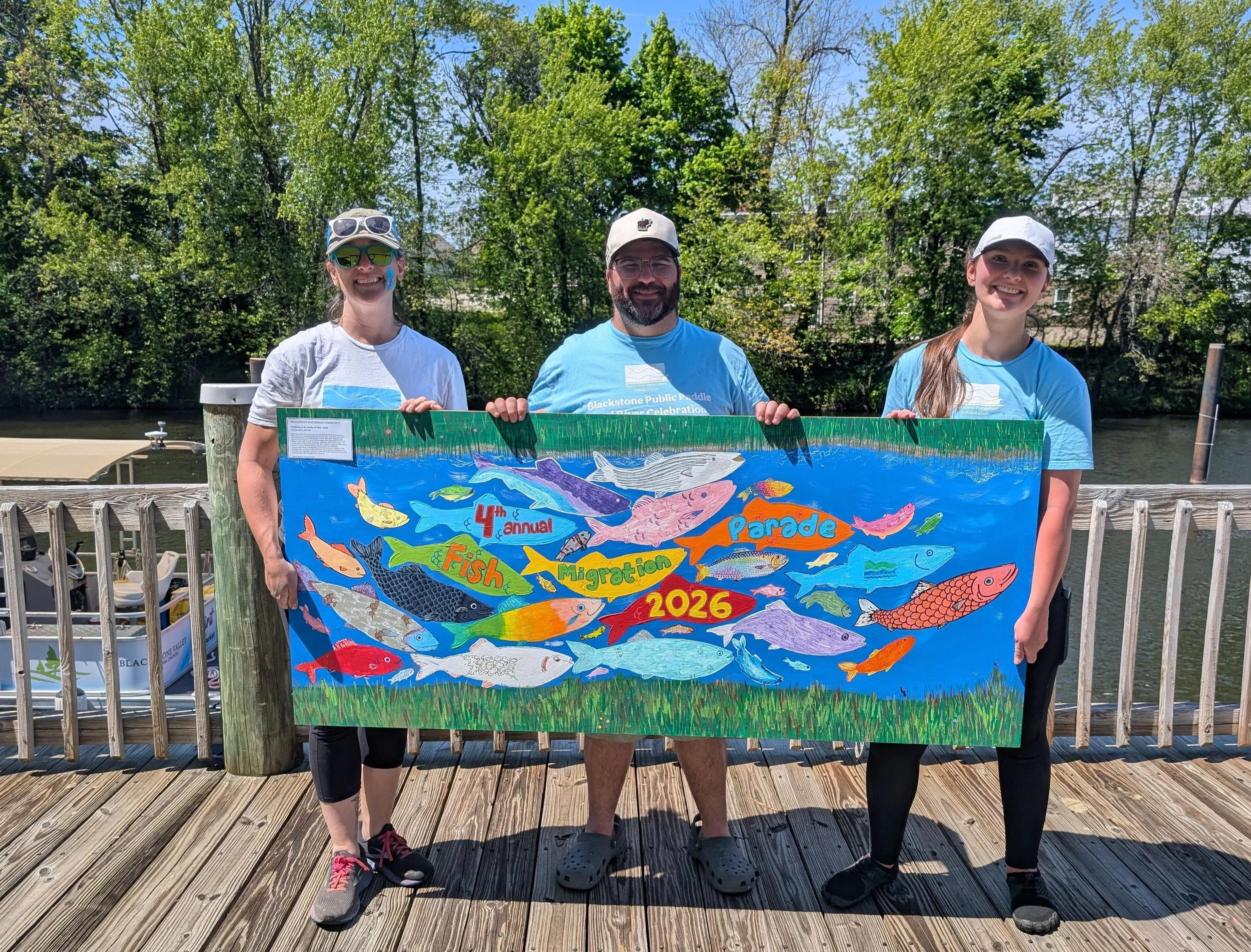 Stefanie, Kevin, and Caleigh hold a wooden board painted with colorful fish. They are standing in front of the river on a wooden walkway, wearing Collaborative shirts and smiling.