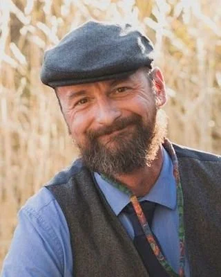 A man with a beard and mustache wearing a beret, a blue shirt, and a vest, smiling outdoors among dry foliage.