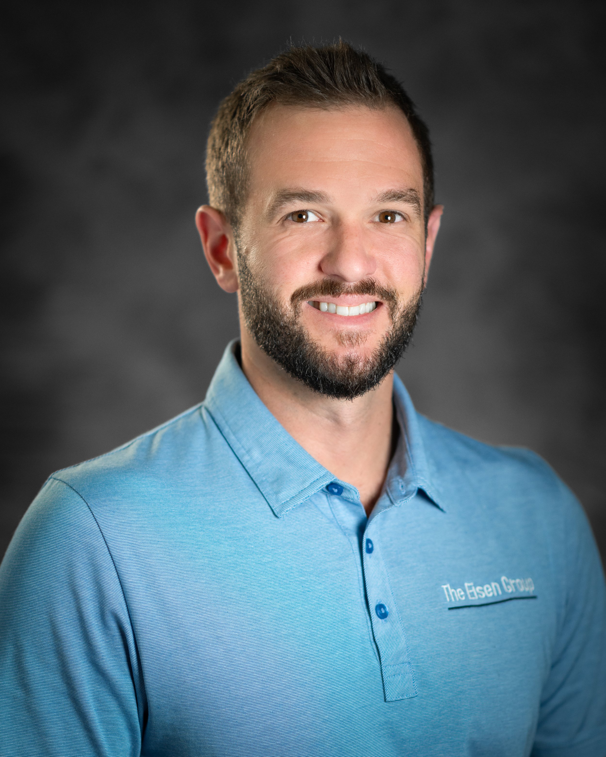 Headshot of a smiling man with short brown hair and beard, wearing a light blue polo shirt with 'The Eisen Group' embroidered on it, against a dark gray background.