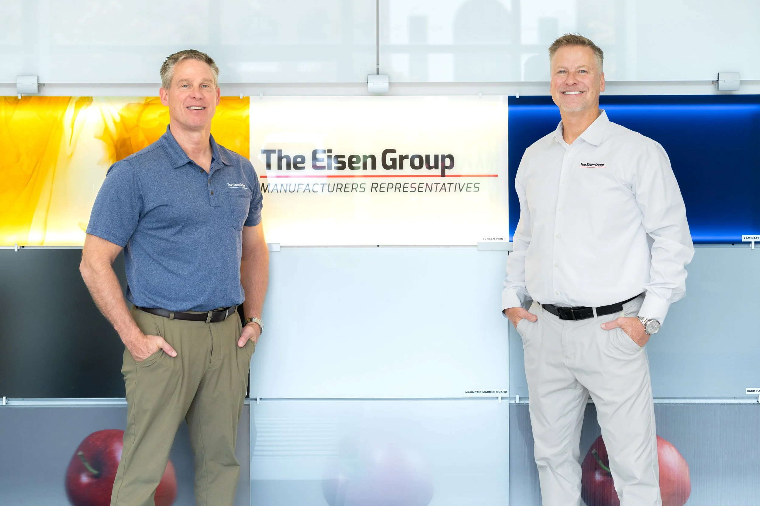 Two men standing in front of a company banner that reads "The Eisen Group Manufactures Representatives" with colorful backgrounds and apple images.