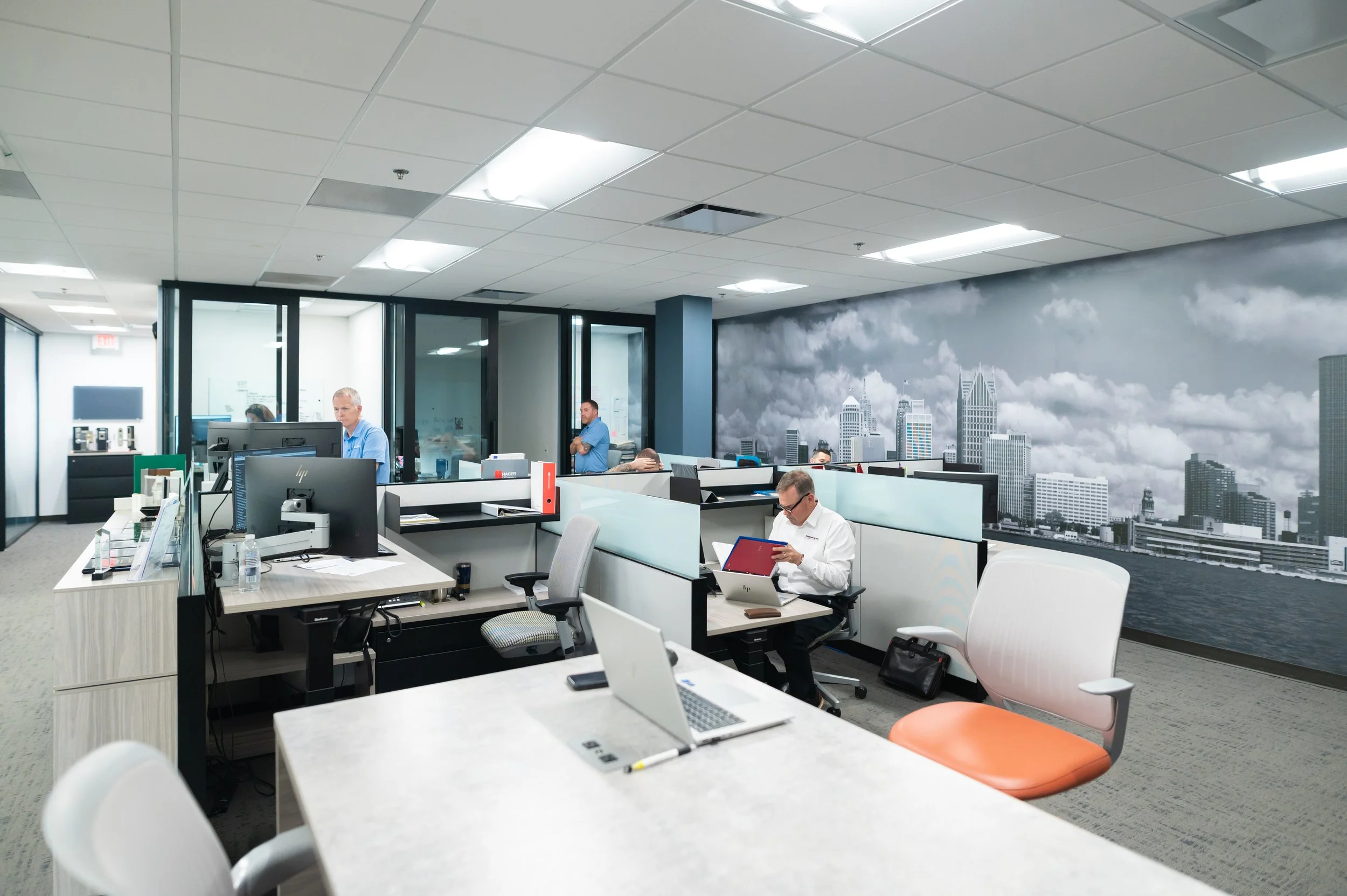 An office with multiple employees working at their desks, some on computers, with a city skyline mural on the wall depicting a cloudy sky and skyscrapers.