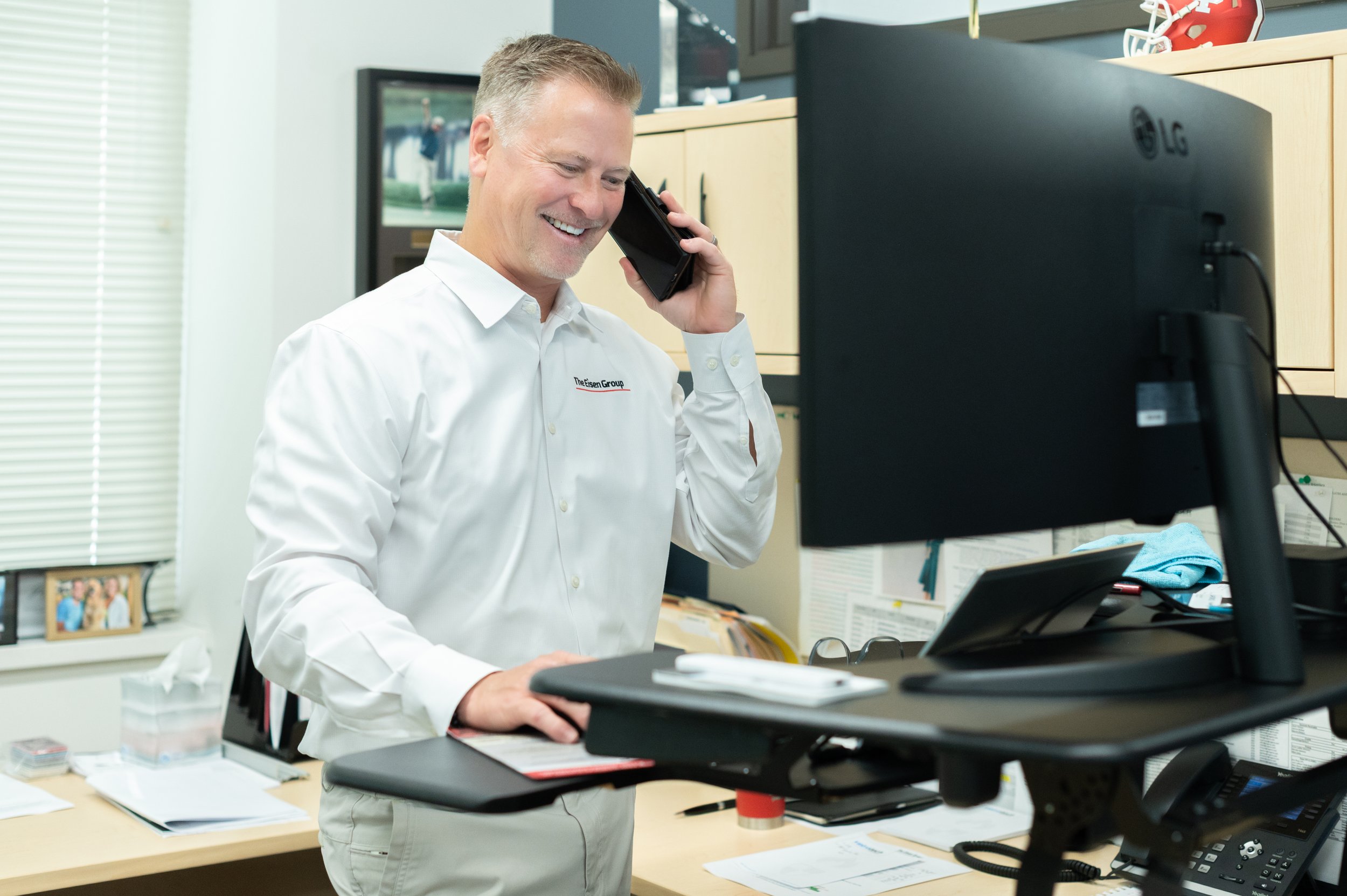 A man in a white shirt smiling and talking on a black phone in an office, standing beside a desk with papers and a computer monitor.