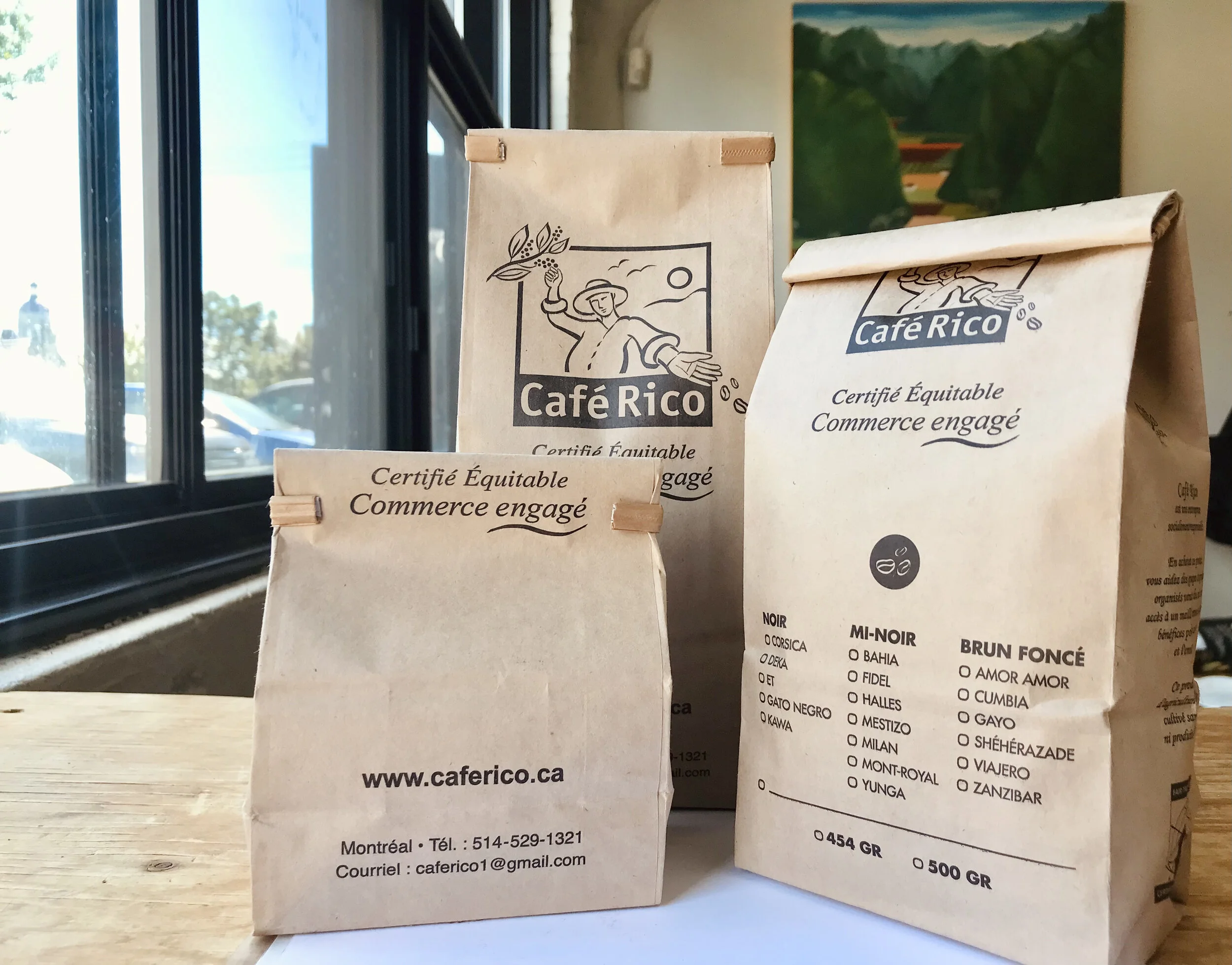 Trois sacs de café de la marque Café Rico posés sur une table en bois, avec une fenêtre derrière.