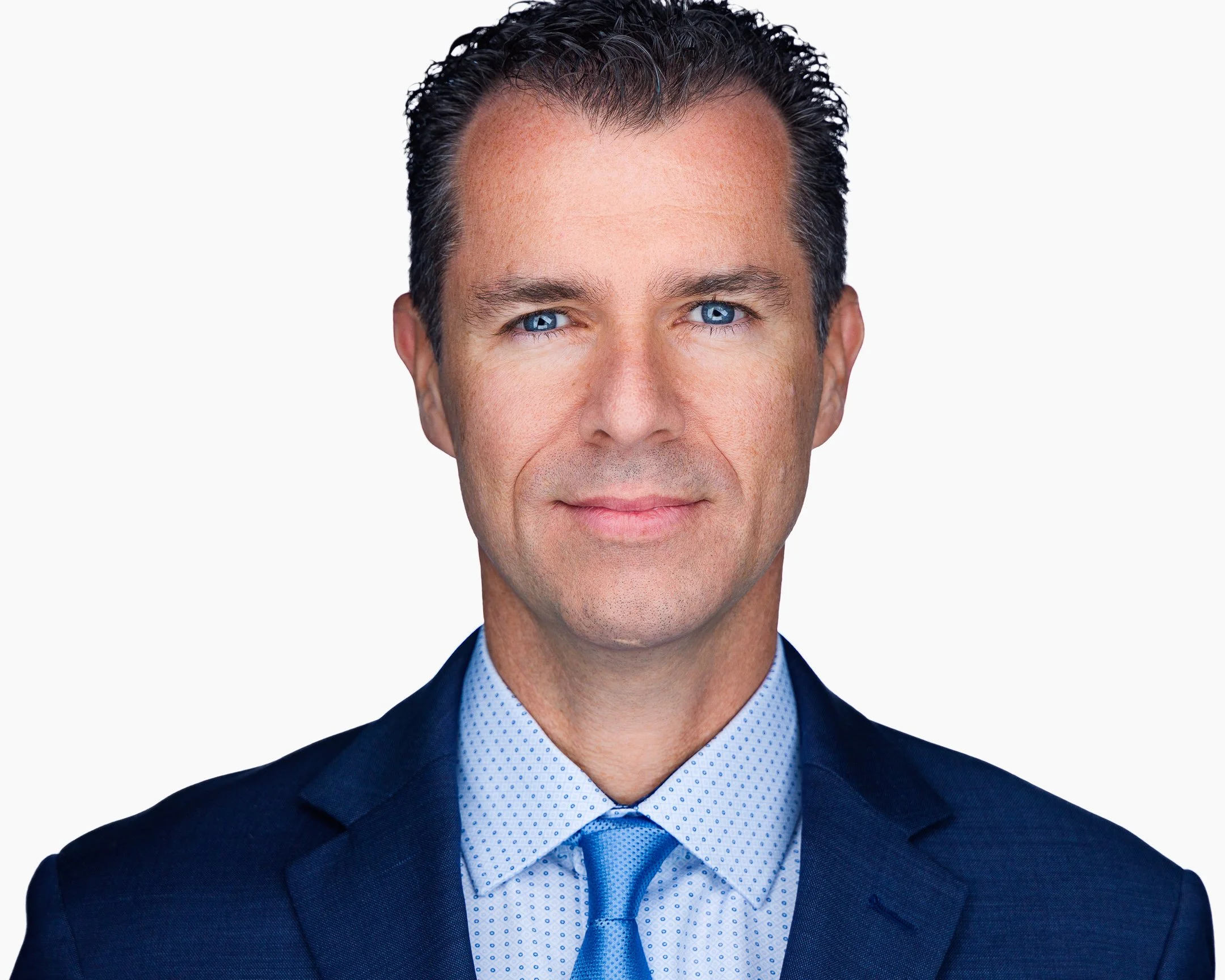 Headshot of a man in a navy blue suit and light blue shirt with a blue tie, smiling with short curly dark hair and blue eyes.