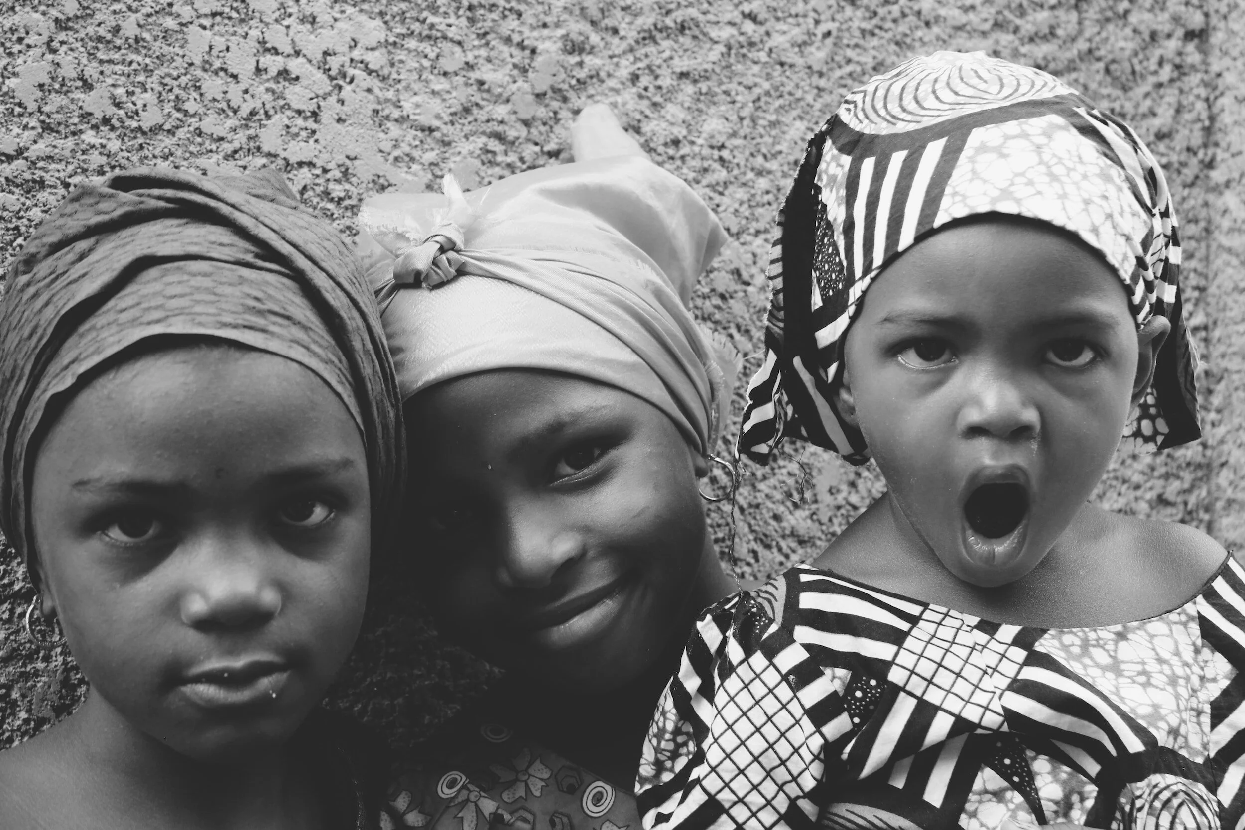 Three young girls with headscarves, looking at the camera outdoors, with one girl on the right yawning.