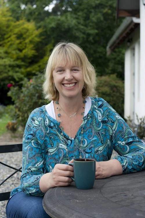Smiling woman in blue patterned shirt holding a mug, seated outdoors at a wooden table with greenery in the background.
