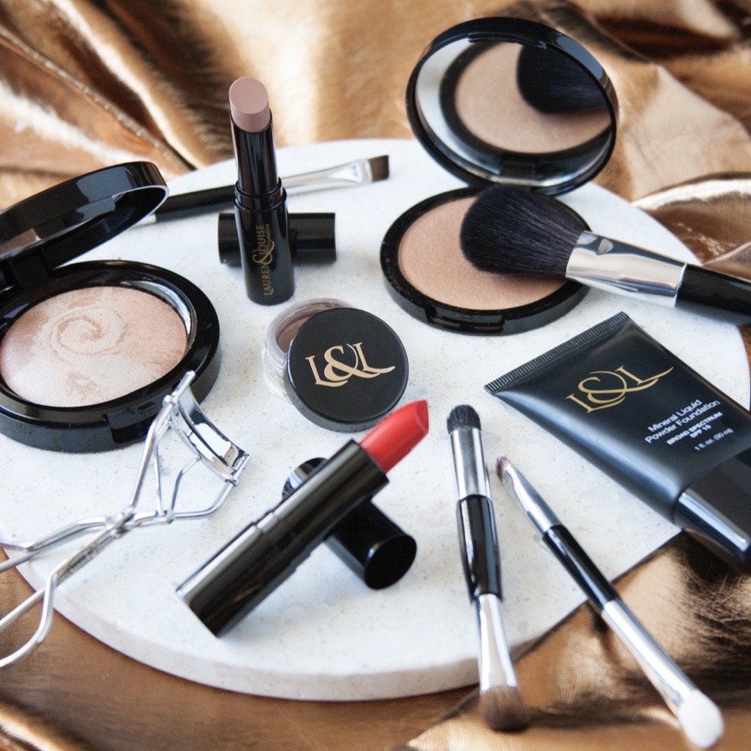 Collection of makeup products including lipstick, blush, brushes, eyelash curler, and foundation on a marble surface.