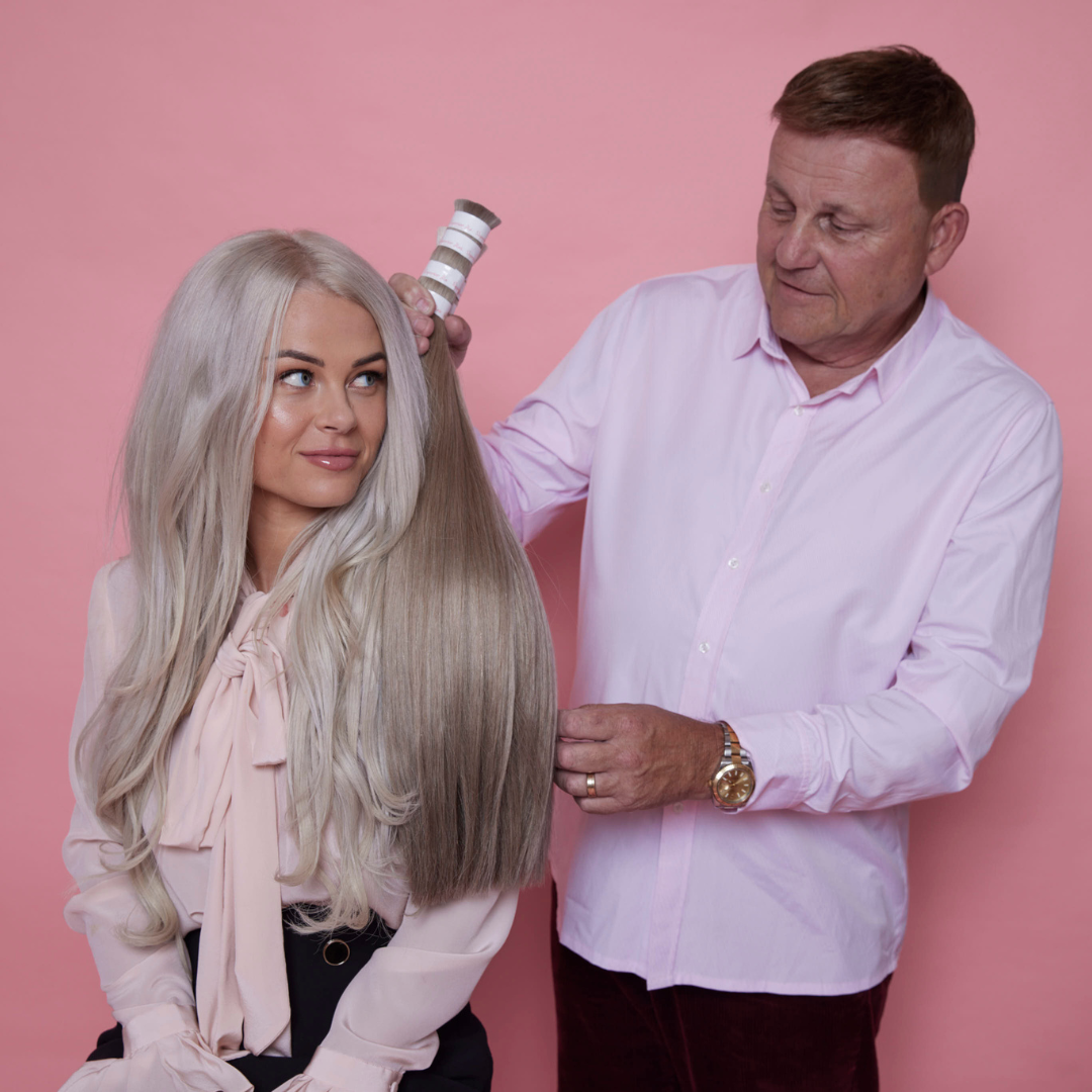 Woman with long blonde hair sitting while a man styles her hair using a brush against a pink background.