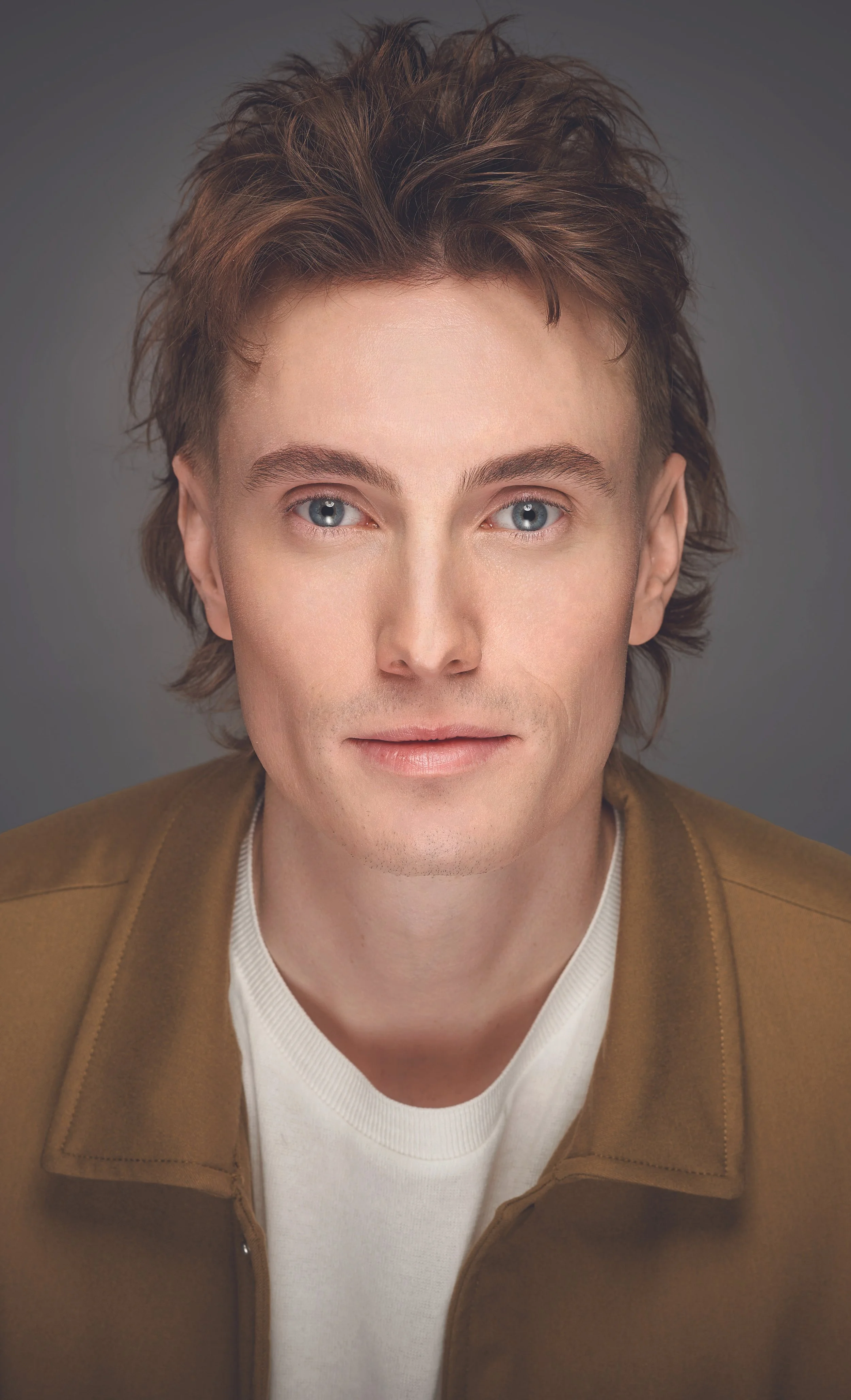 Portrait of a person with short, tousled brown hair, wearing a beige jacket and white T-shirt, against a plain background.
